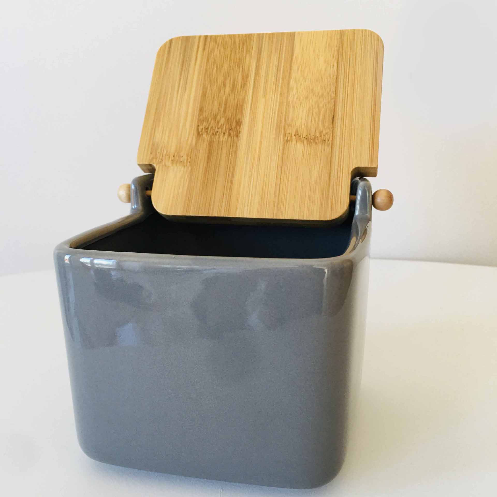 Recipiente cinza com tampa de madeira aberta, sobre superfície branca