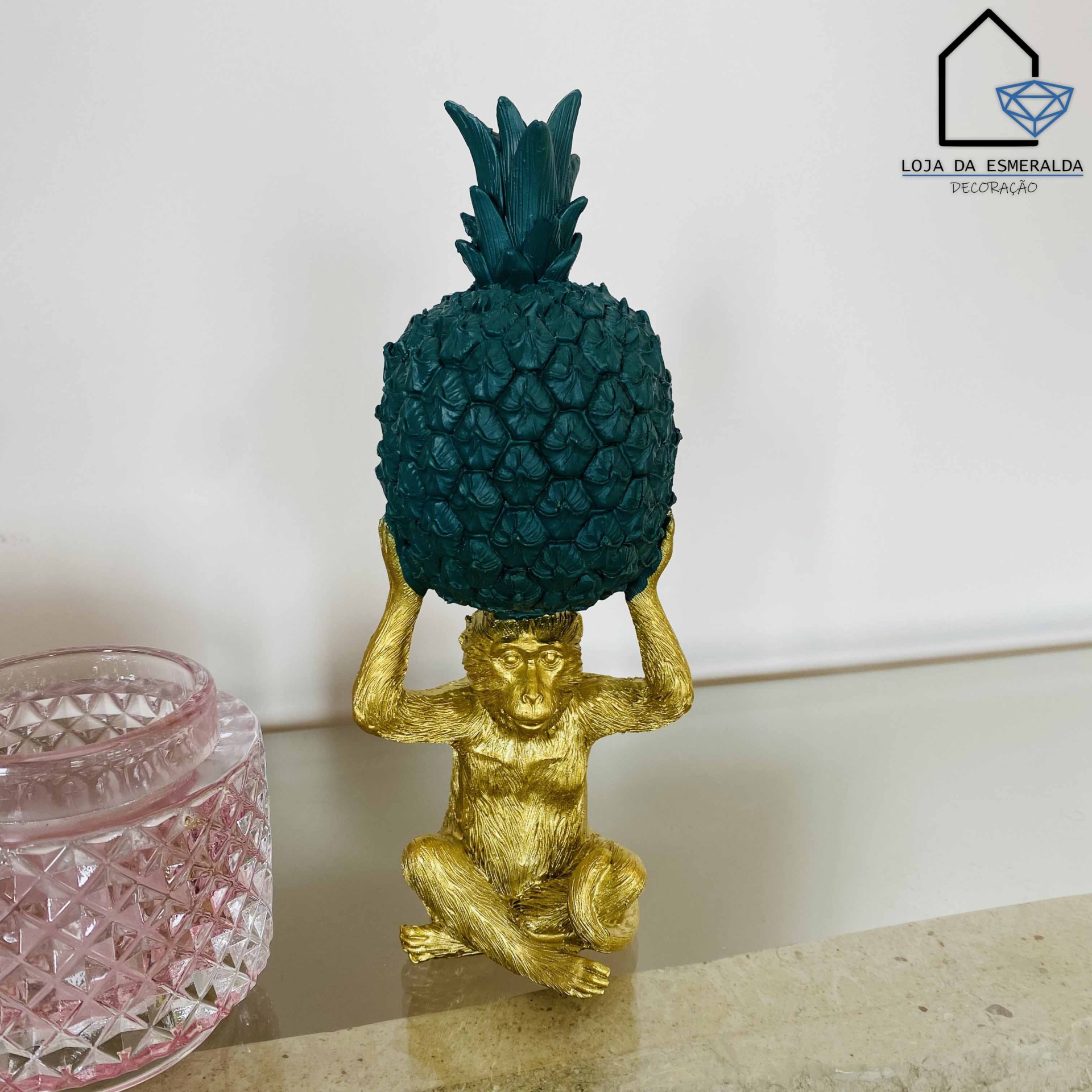 Figura decorativa de macaco dourado com abacaxi verde escuro ao lado de recipiente rosa