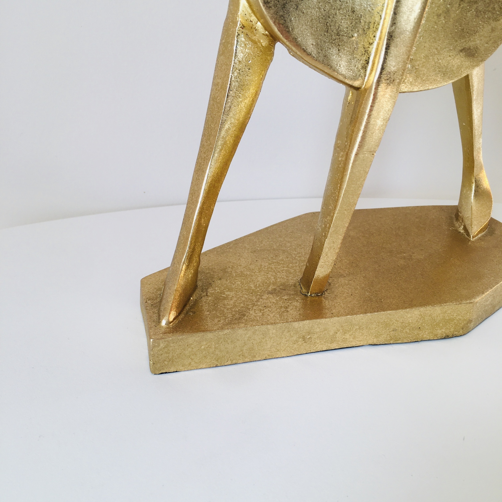 Objeto decorativo dourado com base e três pernas sobre superfície branca