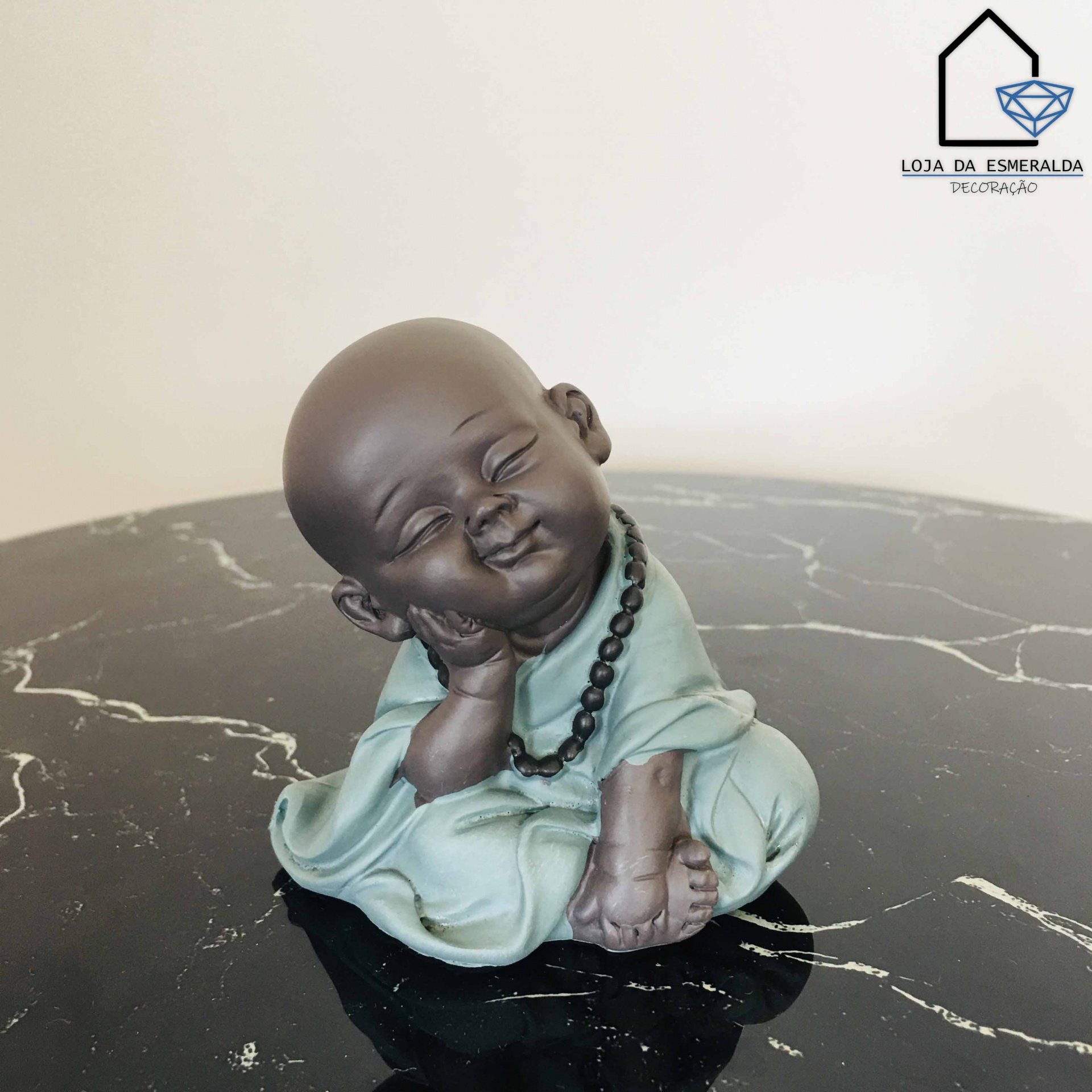 Estátua de monge sorridente sentado com roupa verde clara e colar preto sobre mesa de mármore preto.