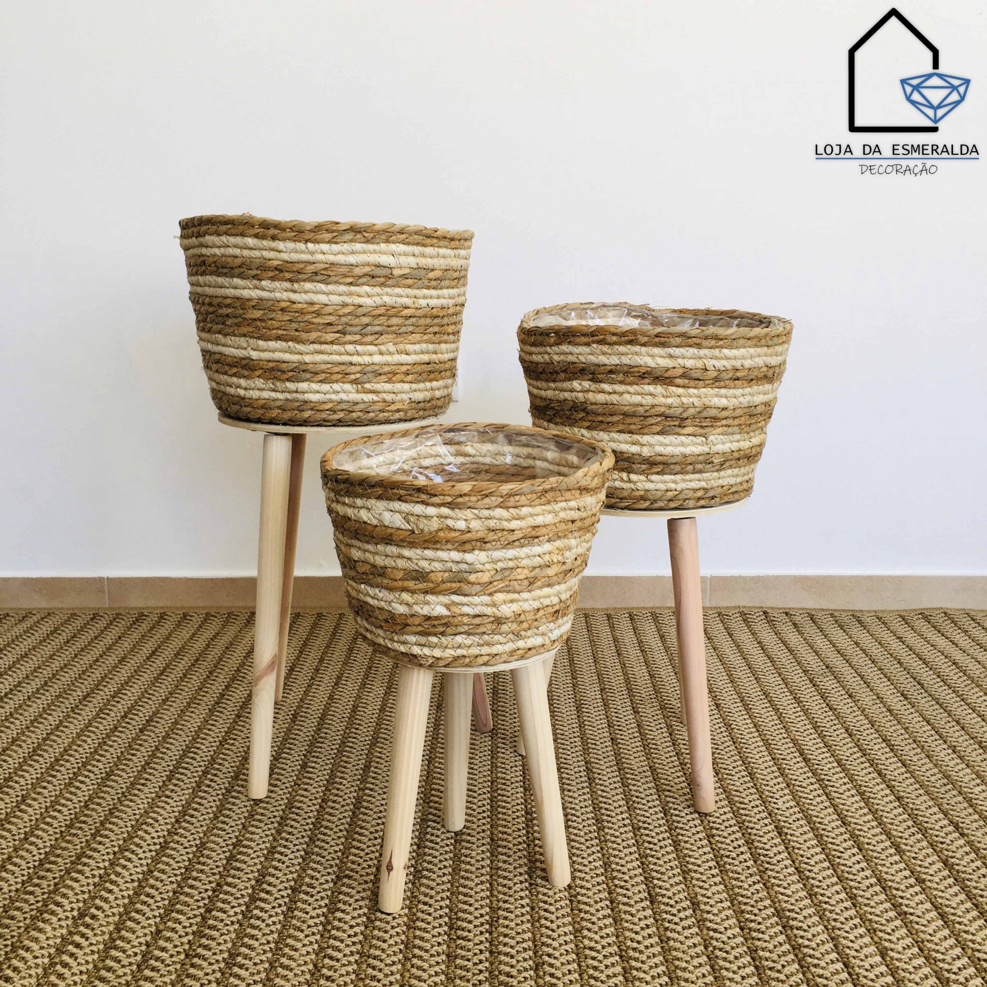 Três cestos em fibras naturais com pernas de madeira clara sobre tapete bege entrelaçado.