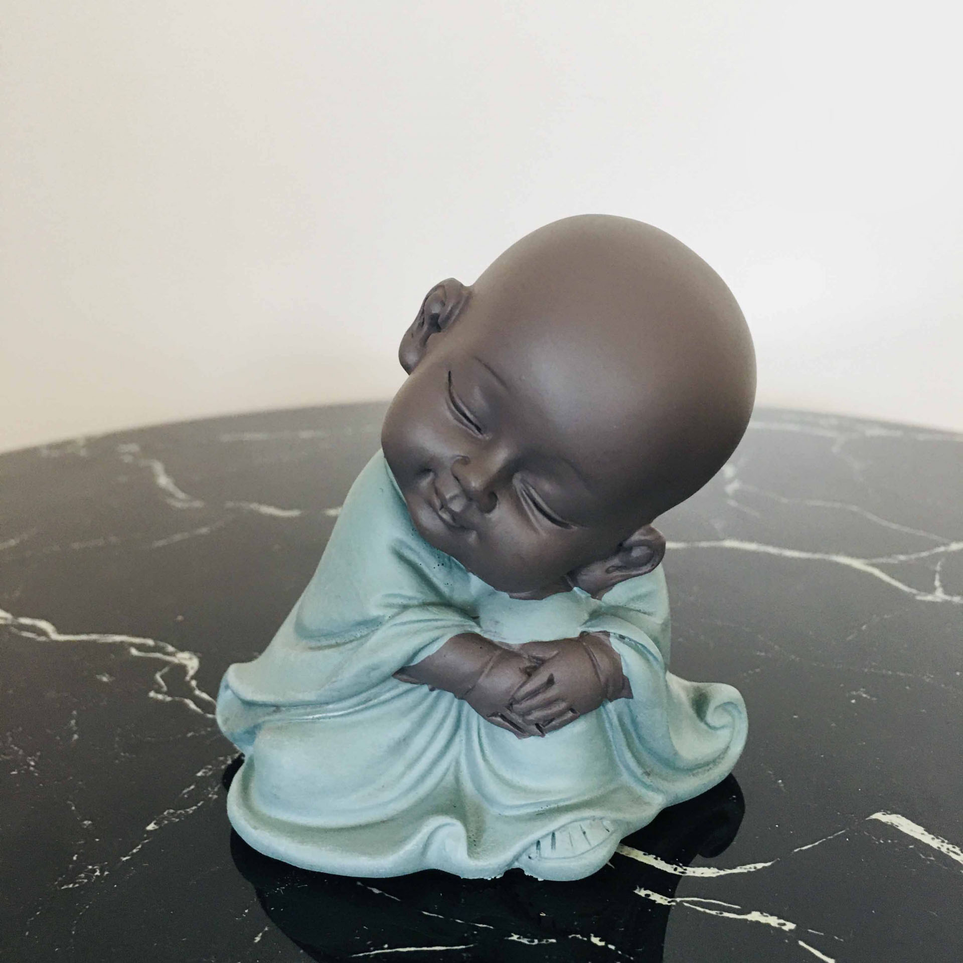 Figura decorativa de um monge bebé sentado com vestes azul esverdeadas numa superfície preta com padrões brancos.