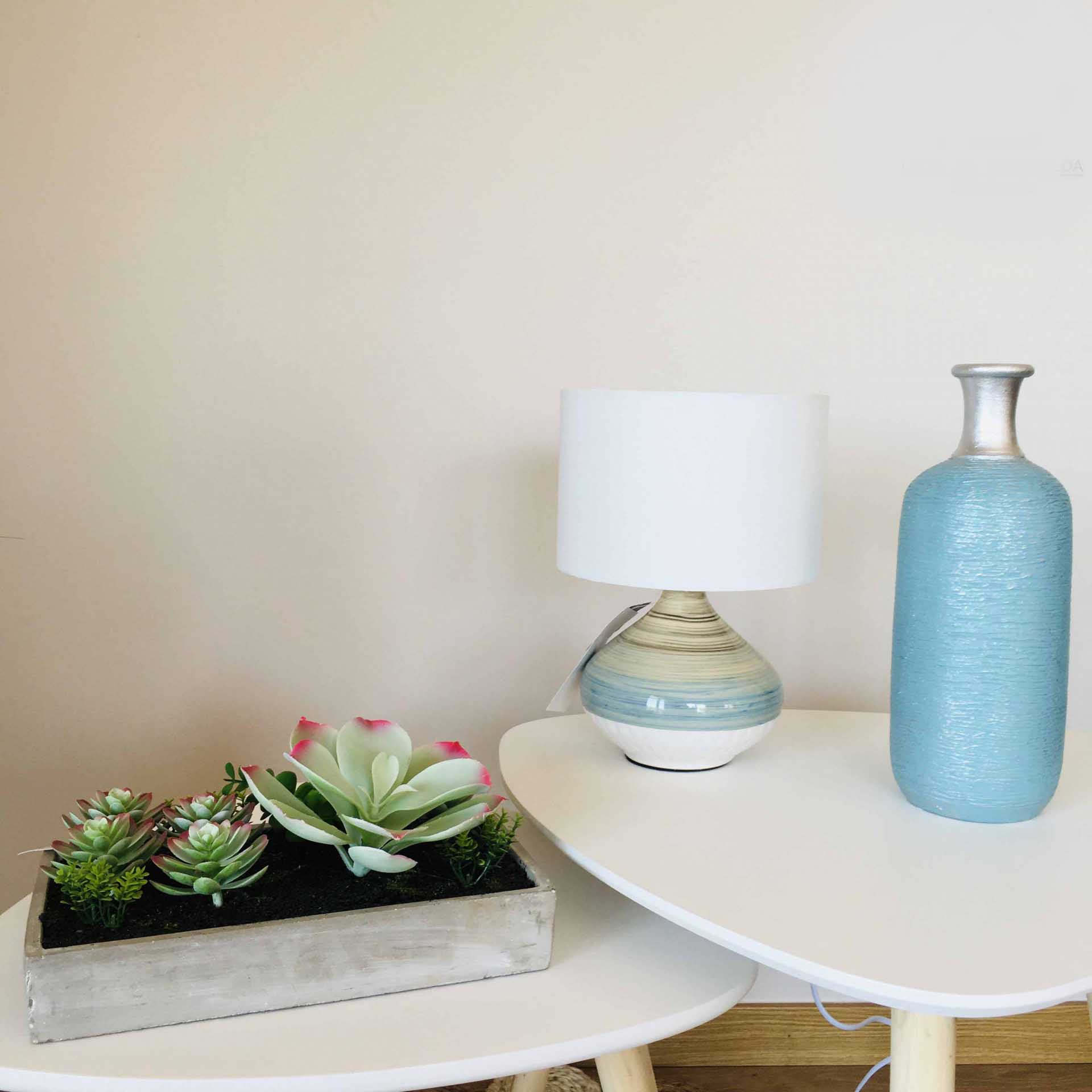 Mesa branca com vaso azul, candeeiro e plantas suculentas artificiais num tabuleiro cinzento