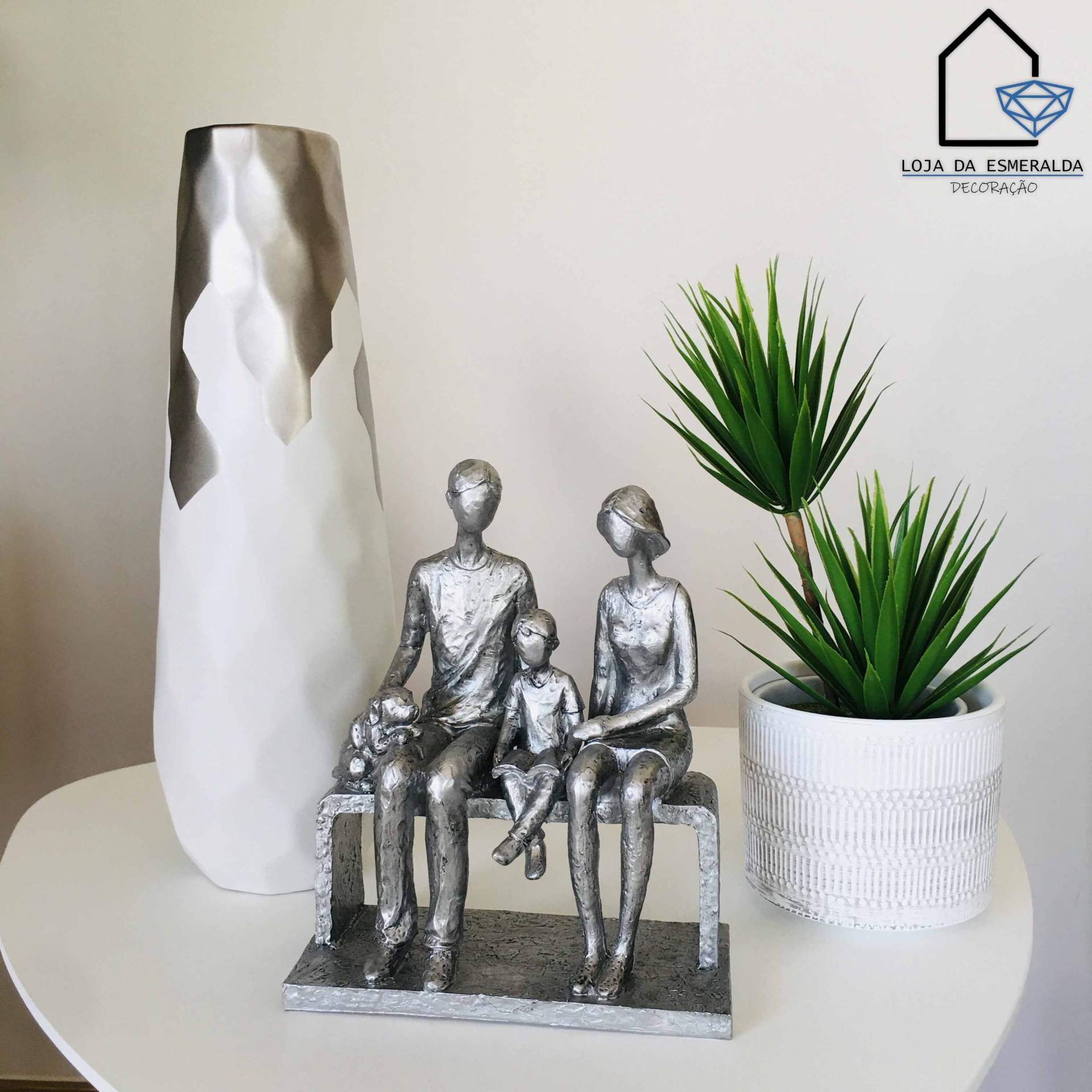 Escultura de família em metal cinzento, vaso branco alto, planta artificial verde em vaso branco, logotipo Loja da Esmeralda Decoração