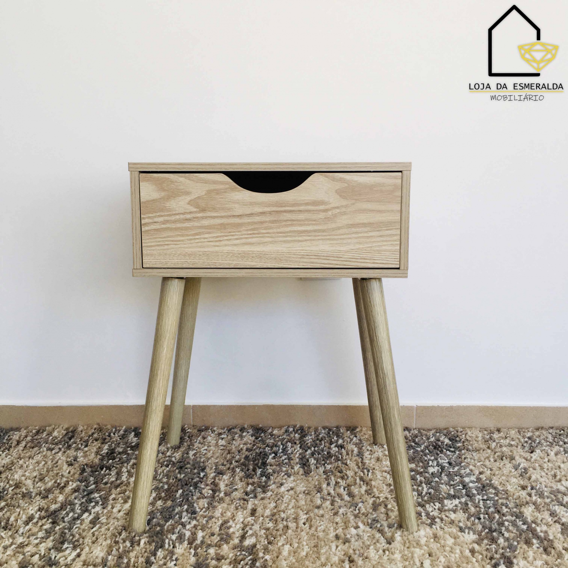 Mesa de cabeceira em madeira clara com gavetão e pernas inclinadas, sobre carpete bege.