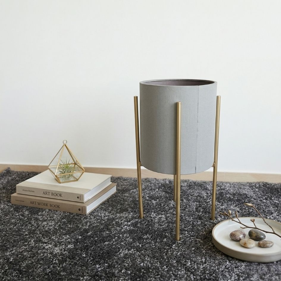 Vaso em tecido cinzento com suporte dourado em tapete cinza, livros e decoração ao lado