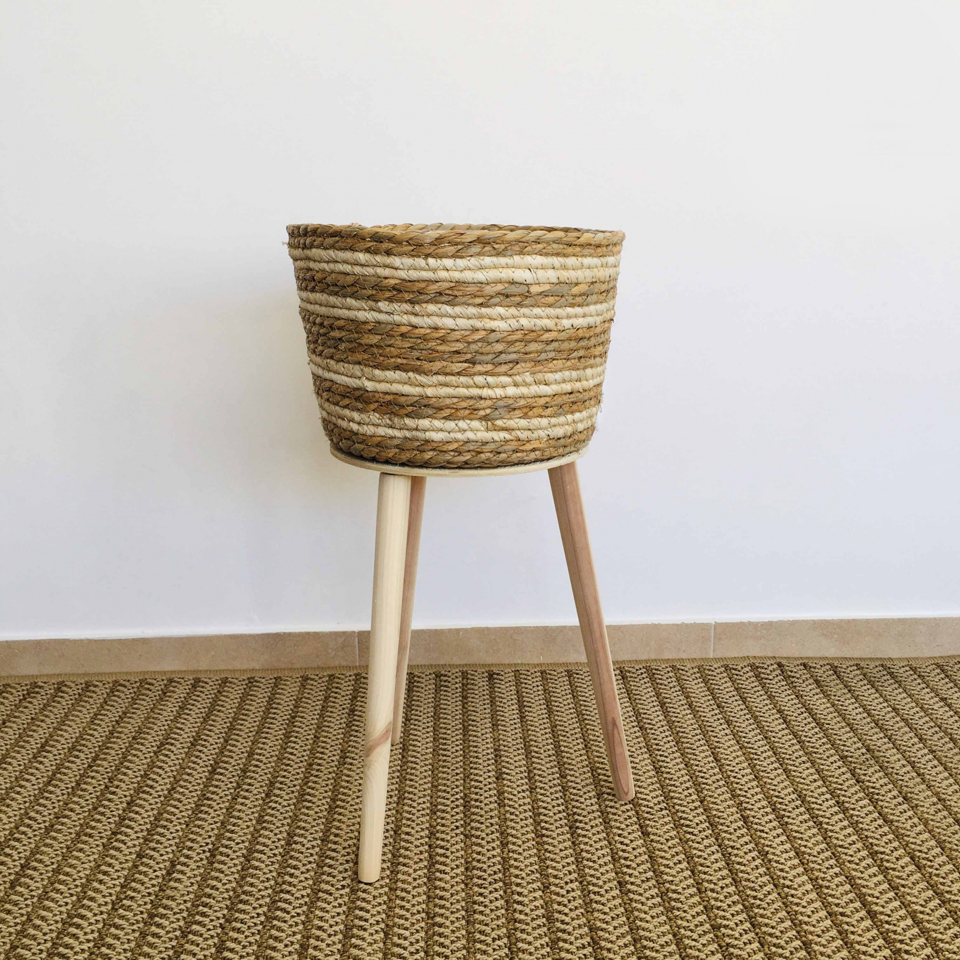 Cesto de palha trançada com pernas de madeira sobre tapete bege
