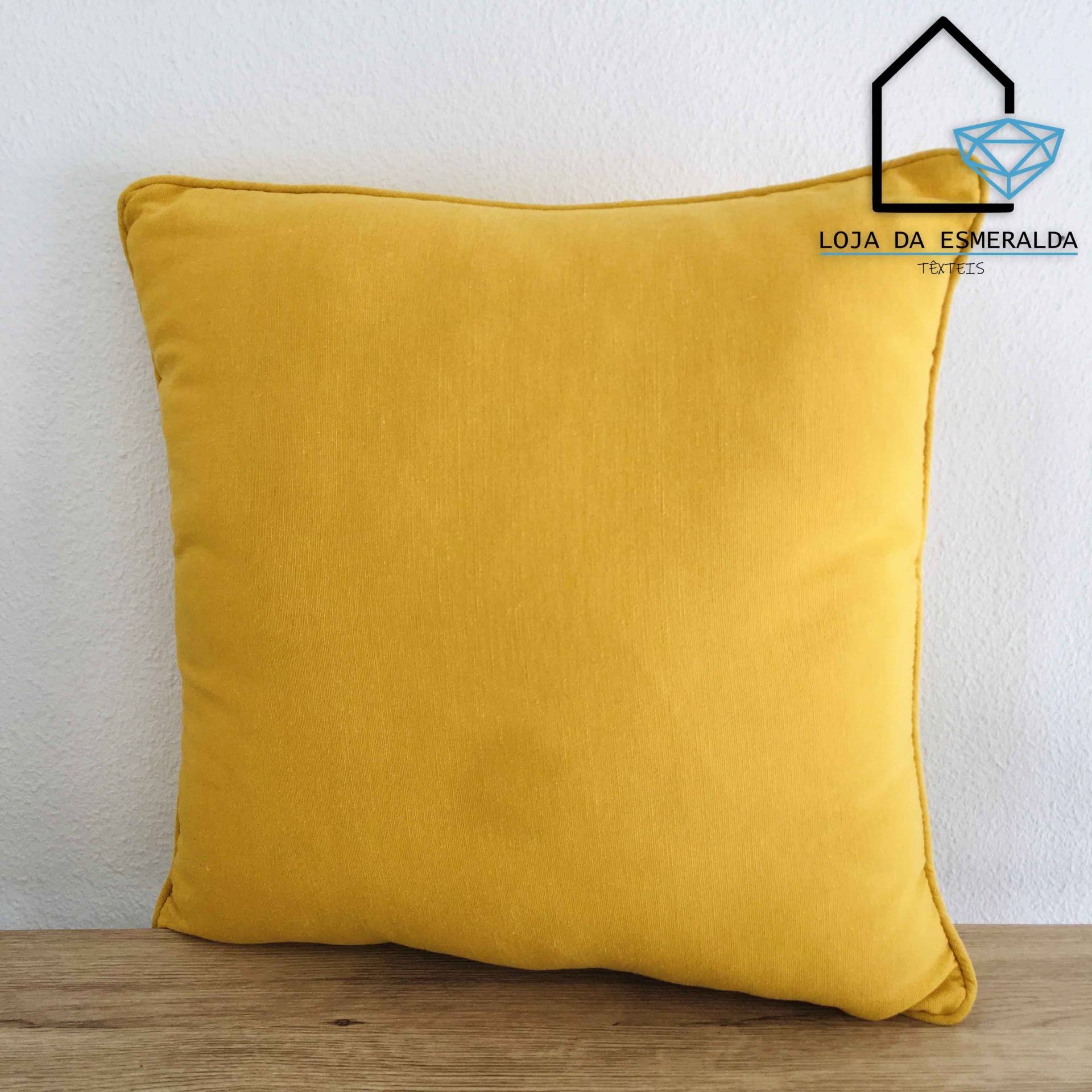 Almofada amarela quadrada sobre superfície de madeira com logótipo da Loja da Esmeralda no canto