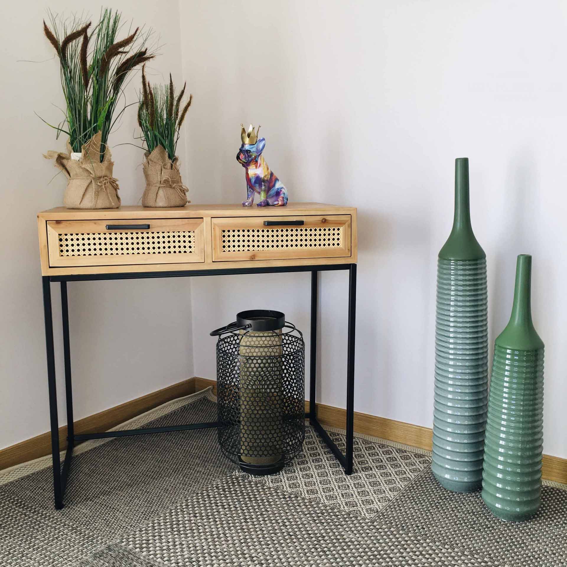 Móvel de madeira clara com gavetas em palhinha e decoração com plantas, estatueta de cão, lanterna e jarras cerâmicas verdes