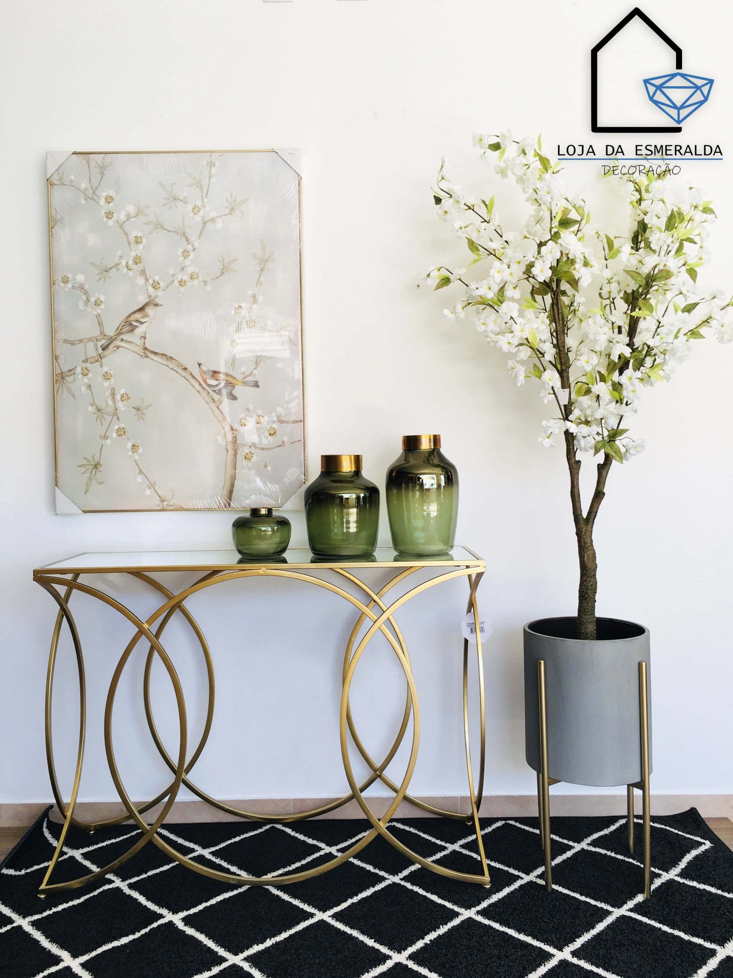 Mesa consola dourada com vasos verdes e quadro floral ao lado de vaso cinzento com flores brancas sobre tapete preto.