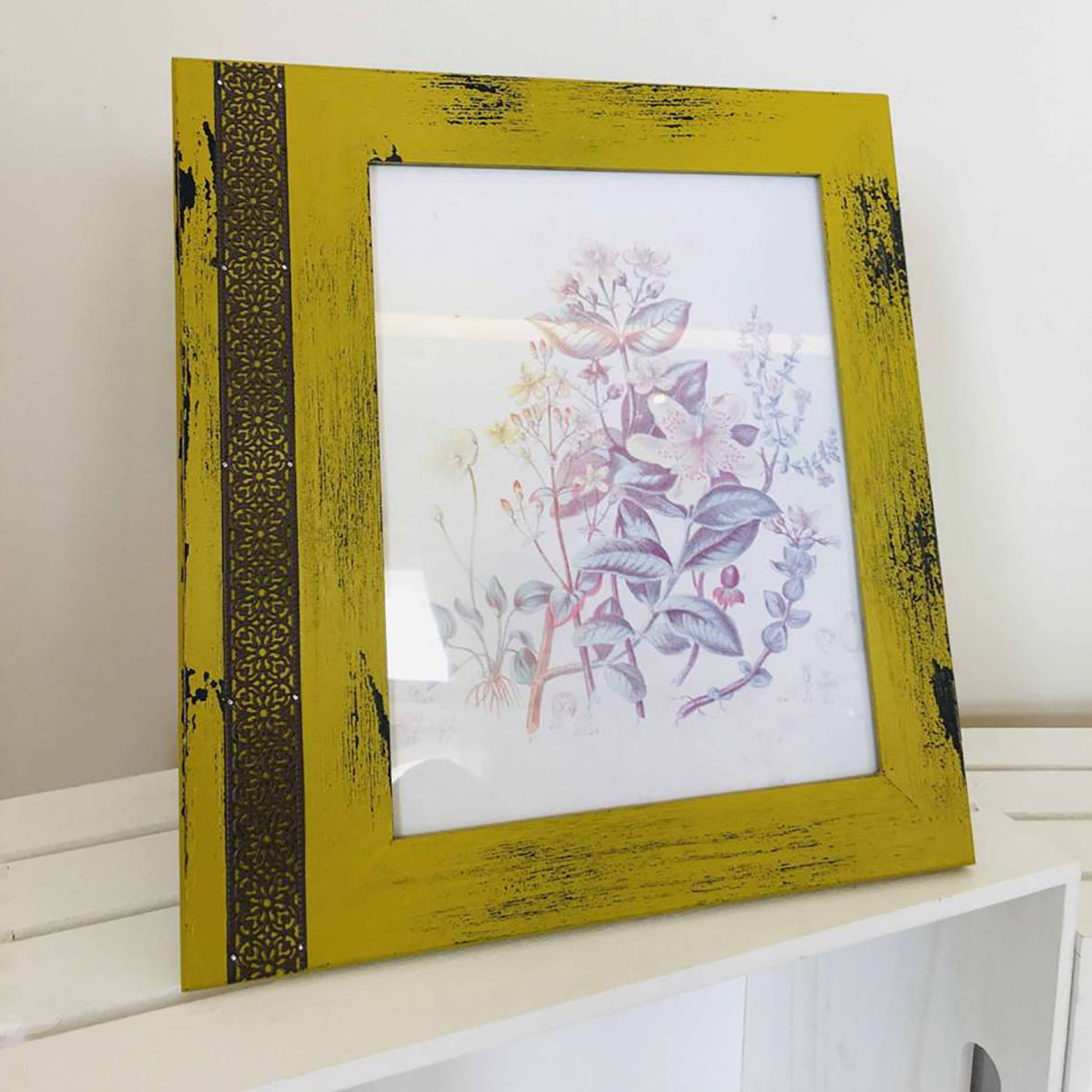 Moldura amarela envelhecida com gravura de plantas colocada num móvel branco