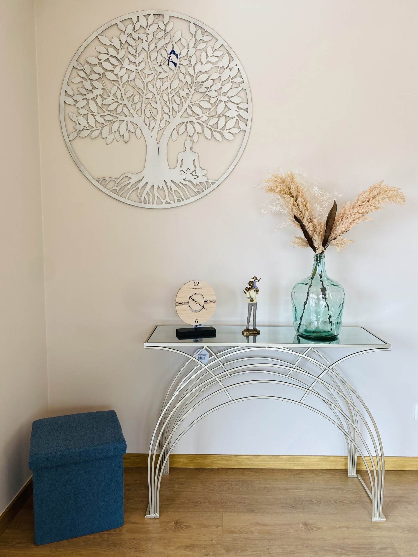 Móvel branco com vidro, relógio de madeira, vaso com ramos secos, pufe azul e decoração de árvore metálica na parede