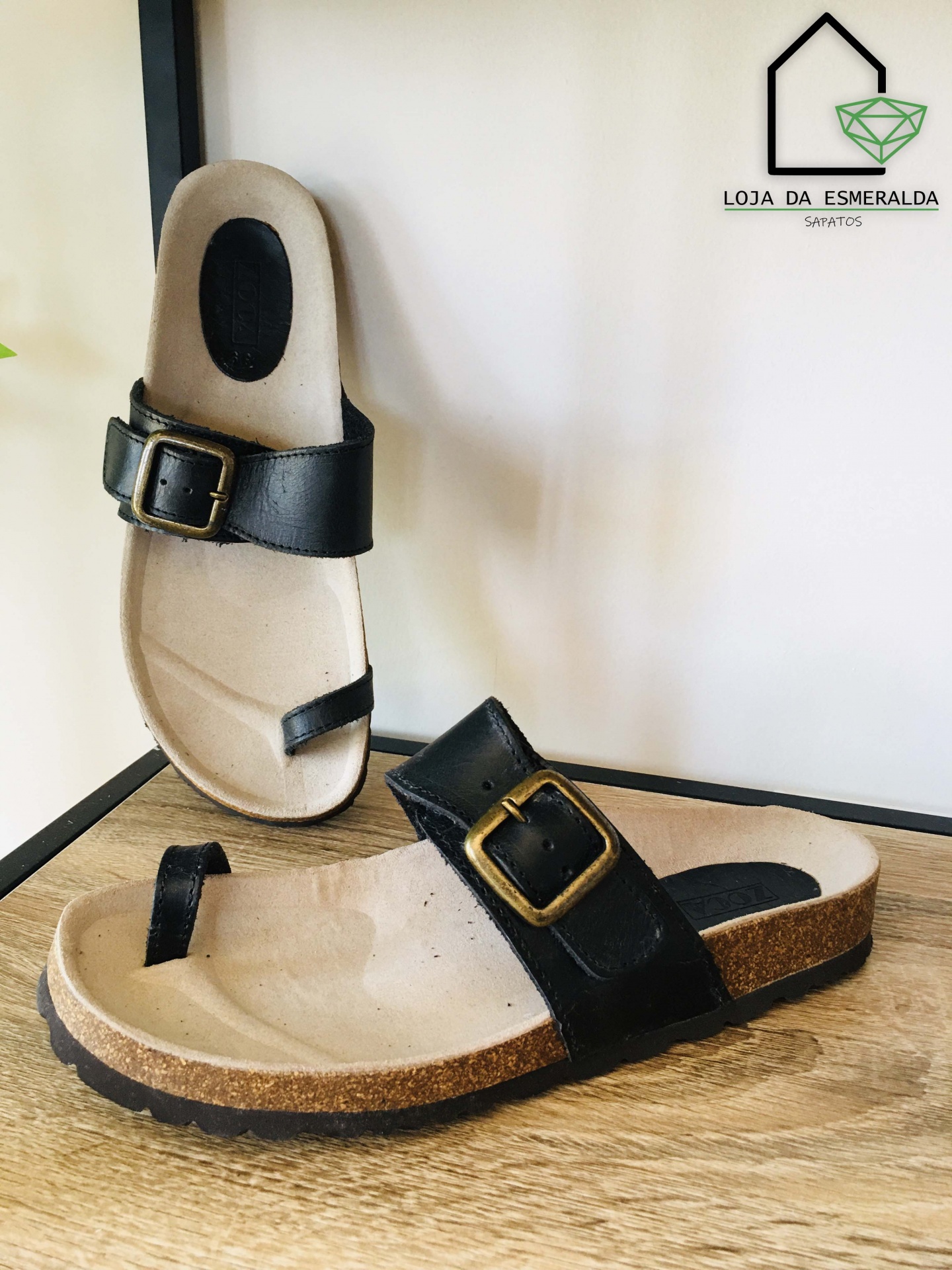 Sandálias castanhas claras com faixa preta e fivela dourada em superfície de madeira com fundo de loja