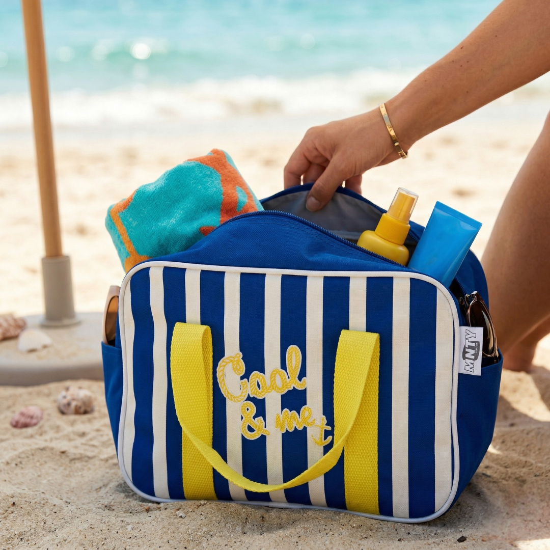 Mala de praia azul às riscas brancas com alças amarelas e bordado, com toalha e protetores solares à beira-mar
