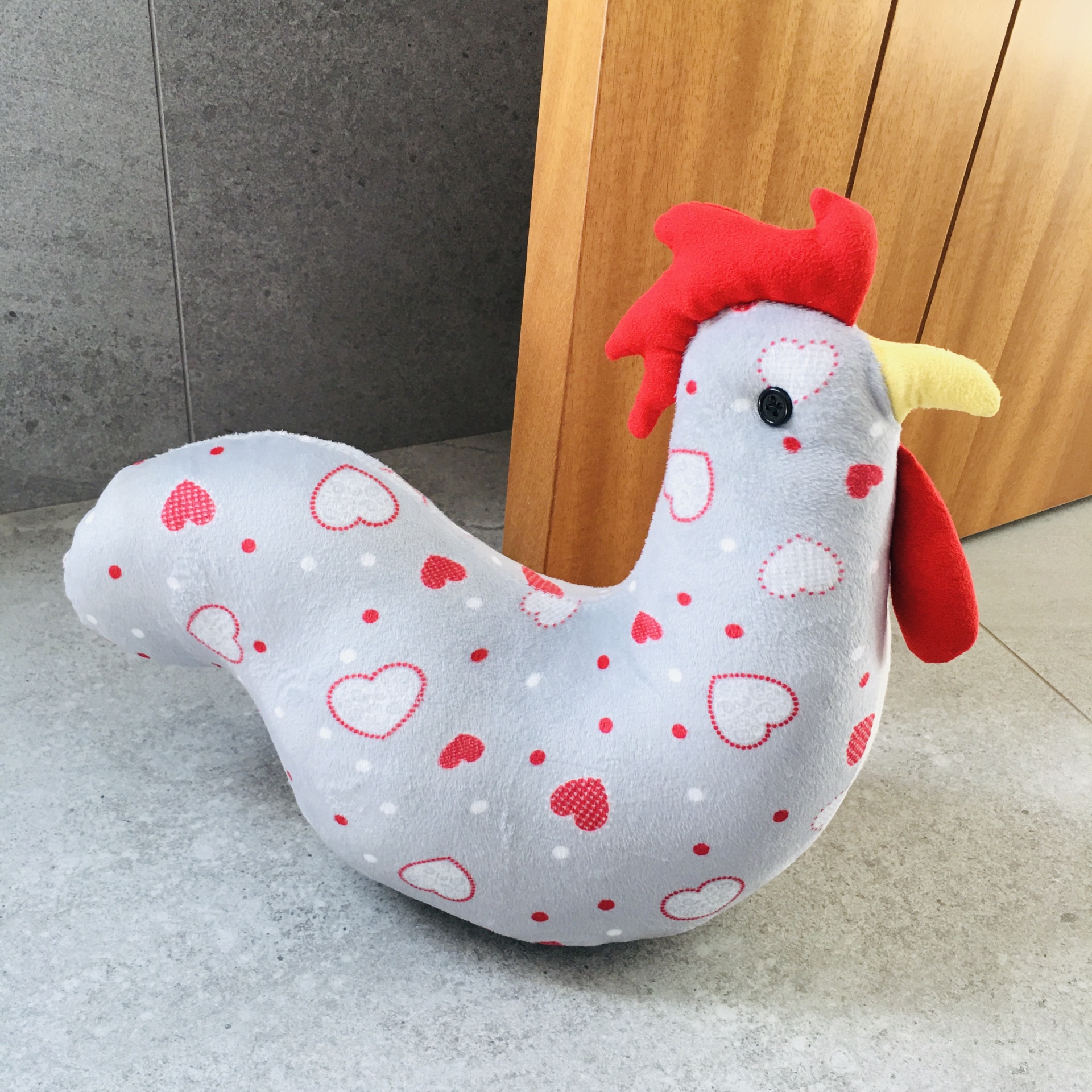 Galo de pelúcia com padrão de corações vermelhos e brancos sobre fundo cinza claro