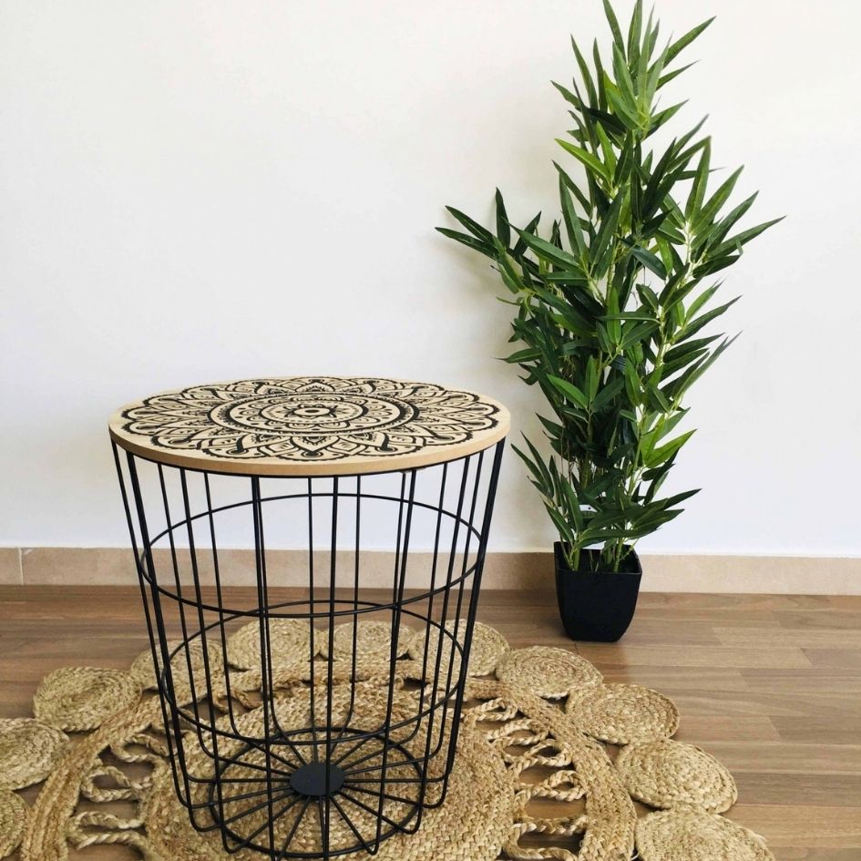 Mesa redonda de madeira com padrão mandala preto e estrutura de metal preto, sobre tapete trançado, ao lado de planta em vaso preto