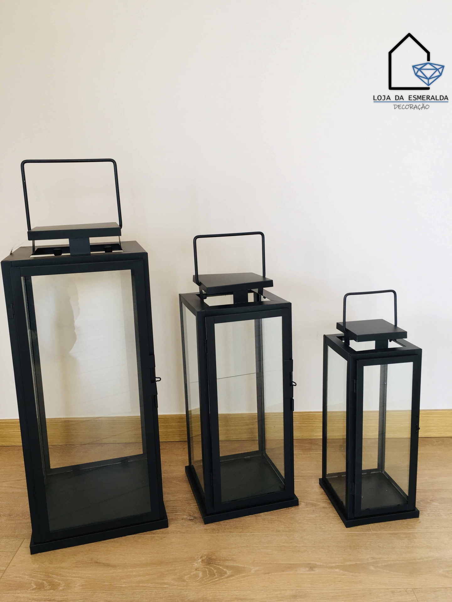Três lanternas decorativas de metal preto e vidro em três tamanhos diferentes sobre chão de madeira