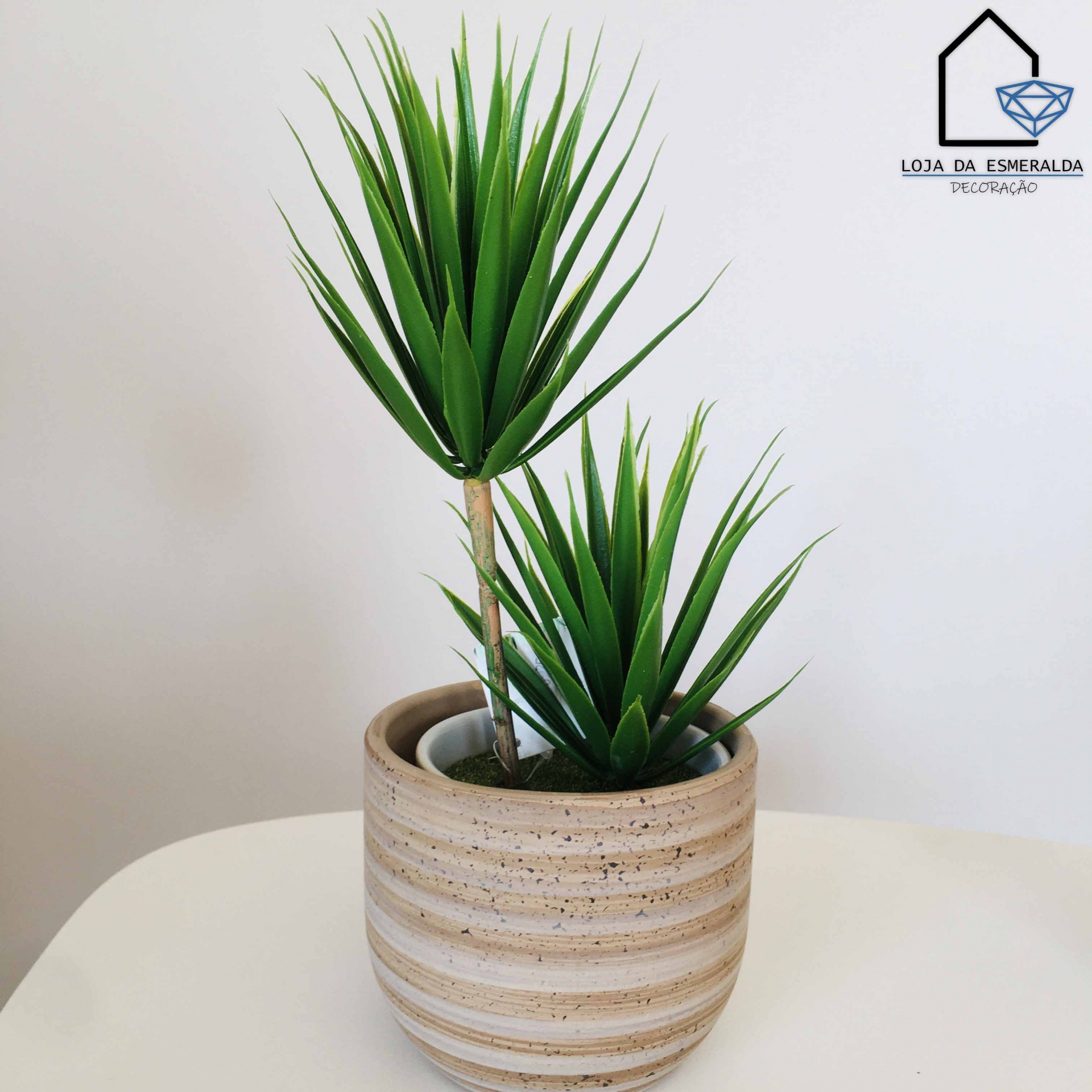 Planta artificial em vaso cerâmico bege com riscas brancas sobre superfície branca