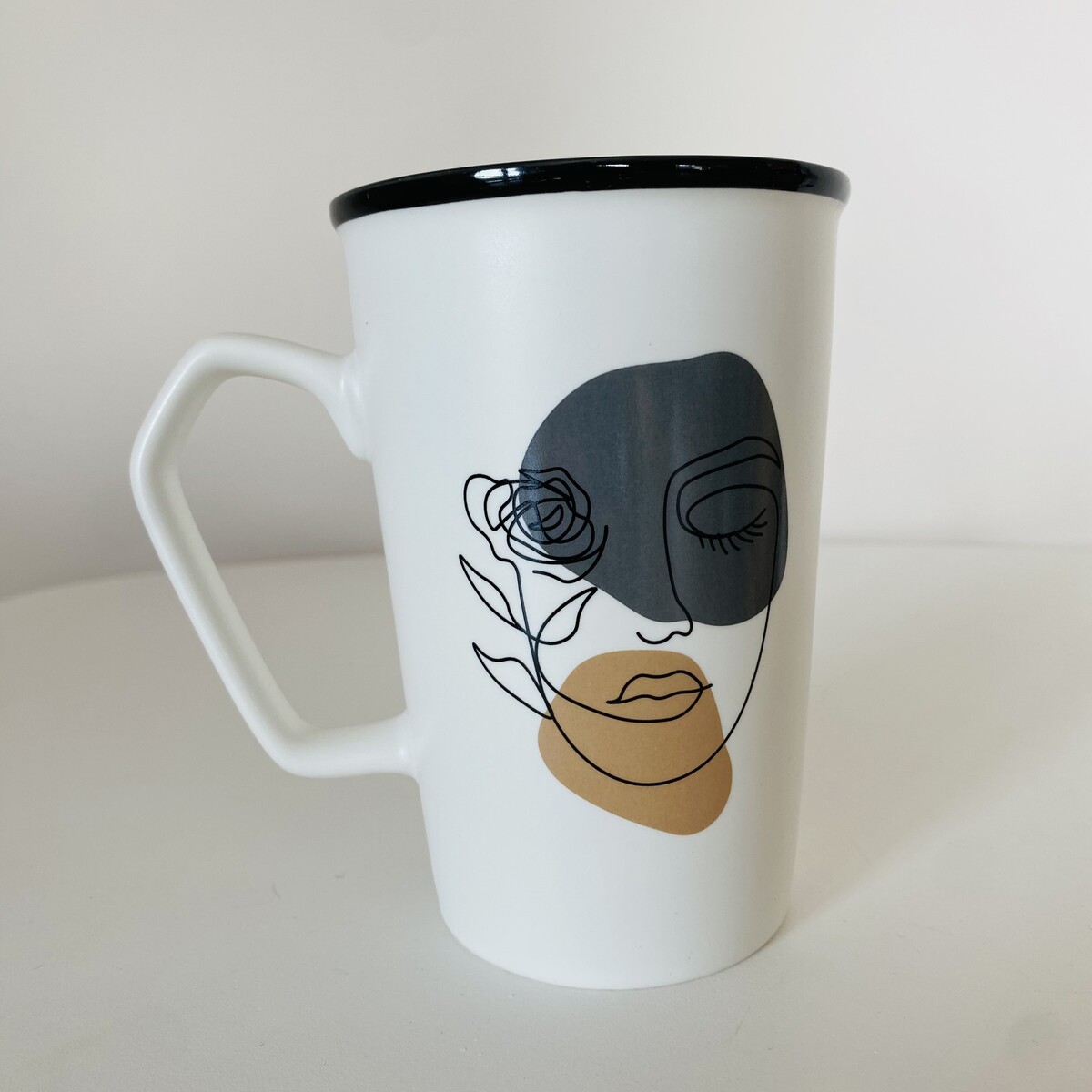 Caneca branca com desenho de rosto feminino em linha contínua com detalhes em cinzento e bege.