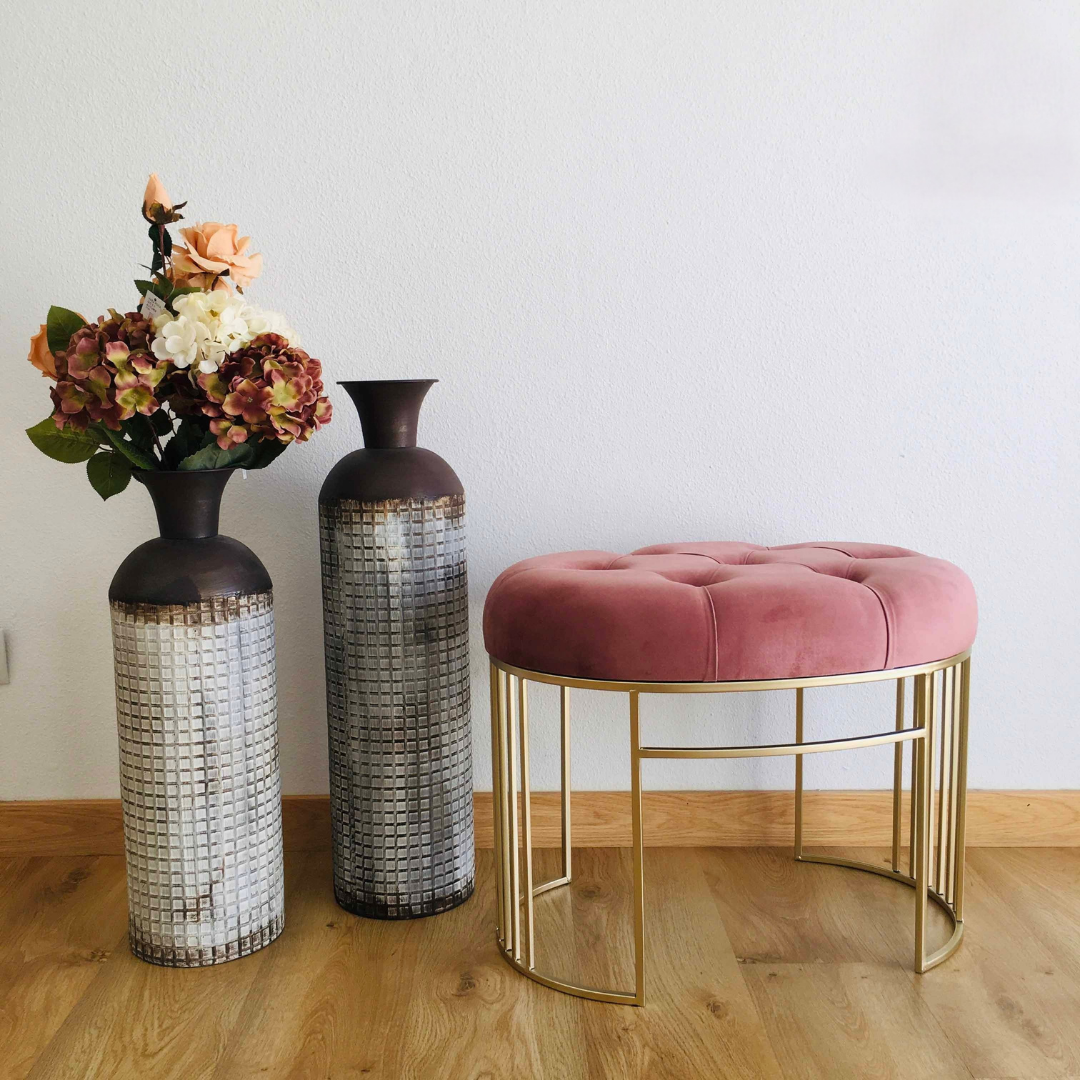Banqueta rosa em veludo com base dourada ao lado de dois vasos cerâmicos altos com flores artificiais