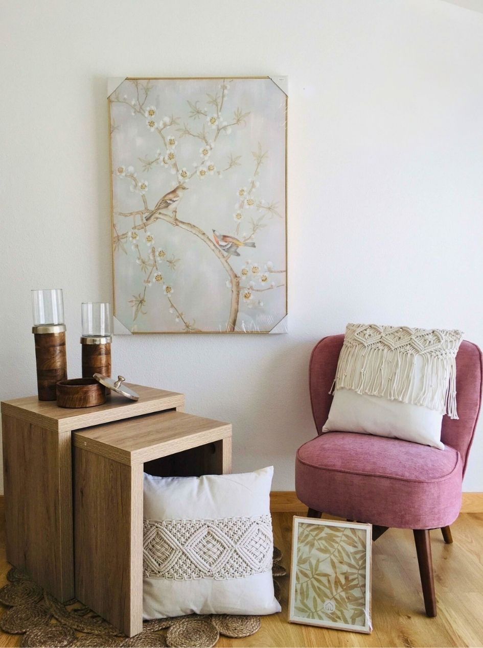 Conjunto de móveis em madeira clara, almofadas macramé brancas, cadeira rosa, copos com suporte de madeira e vidro, caixa de madeira, quadros decorativos com pássaros e folhas.
