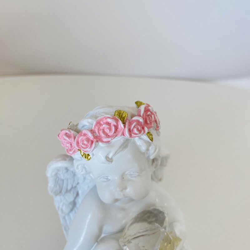 Estátua branca de anjo com coroa de flores rosas e asas, segurando objeto translúcido