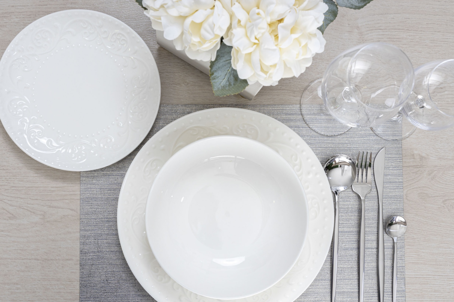 Conjunto de louça branca com padrão floral, talheres em inox, copos de vidro e vaso de flores brancas sobre mesa com individual cinzento