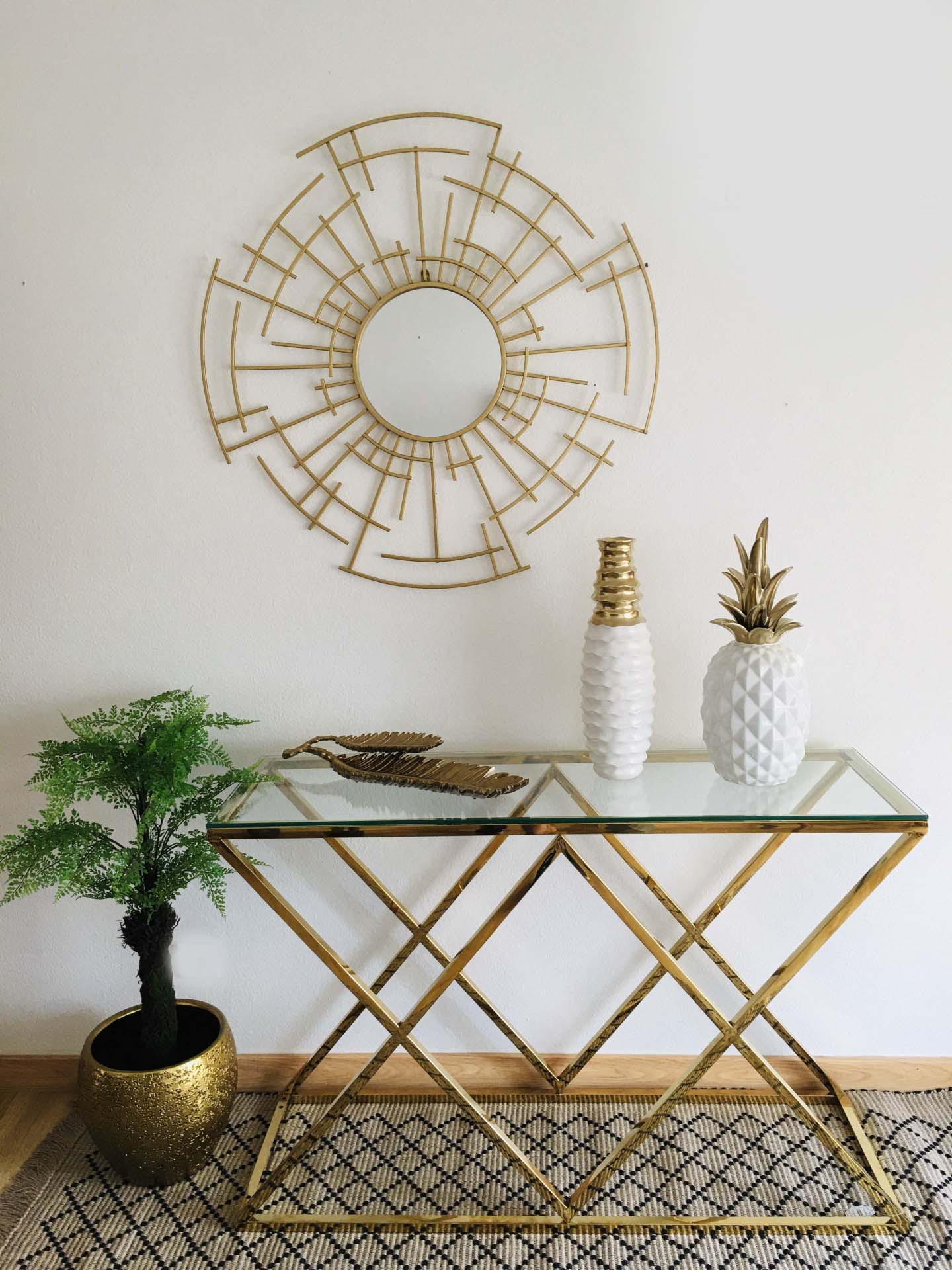 Mesa de vidro com estrutura dourada e decoração com vaso, jarro e planta junto a tapete e espelho redondo dourado na parede