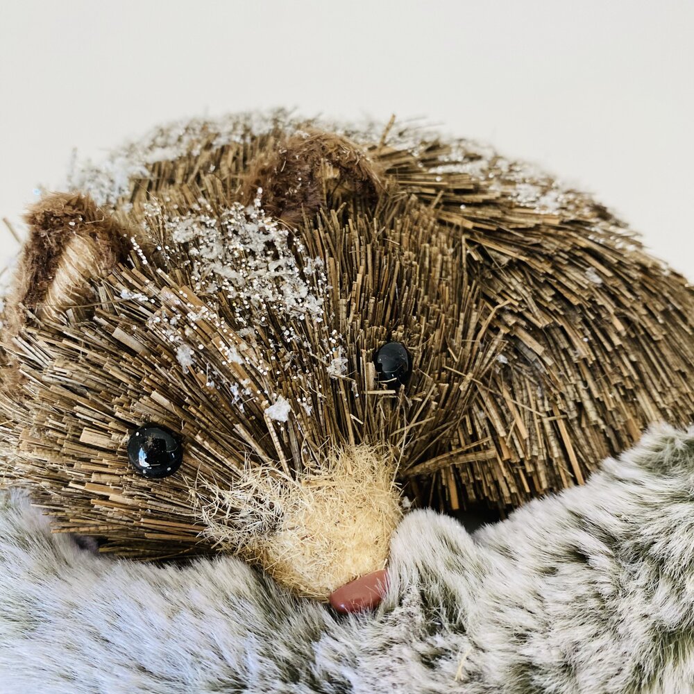 Objeto decorativo em forma de texugo com palha e olhos pretos sobre tecido de pelo