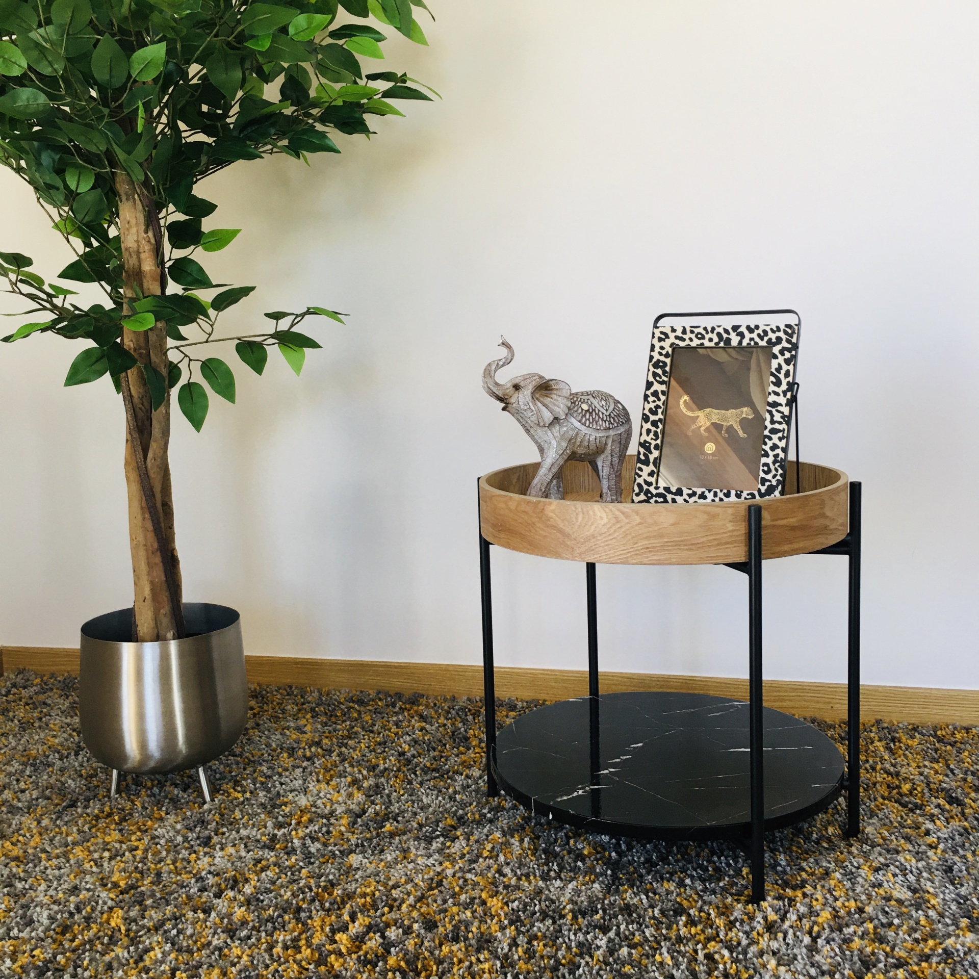Mesa redonda pequena com tampo de madeira clara, base preta, estatueta de elefante e porta-retratos, ao lado de planta em vaso prateado sob tapete.