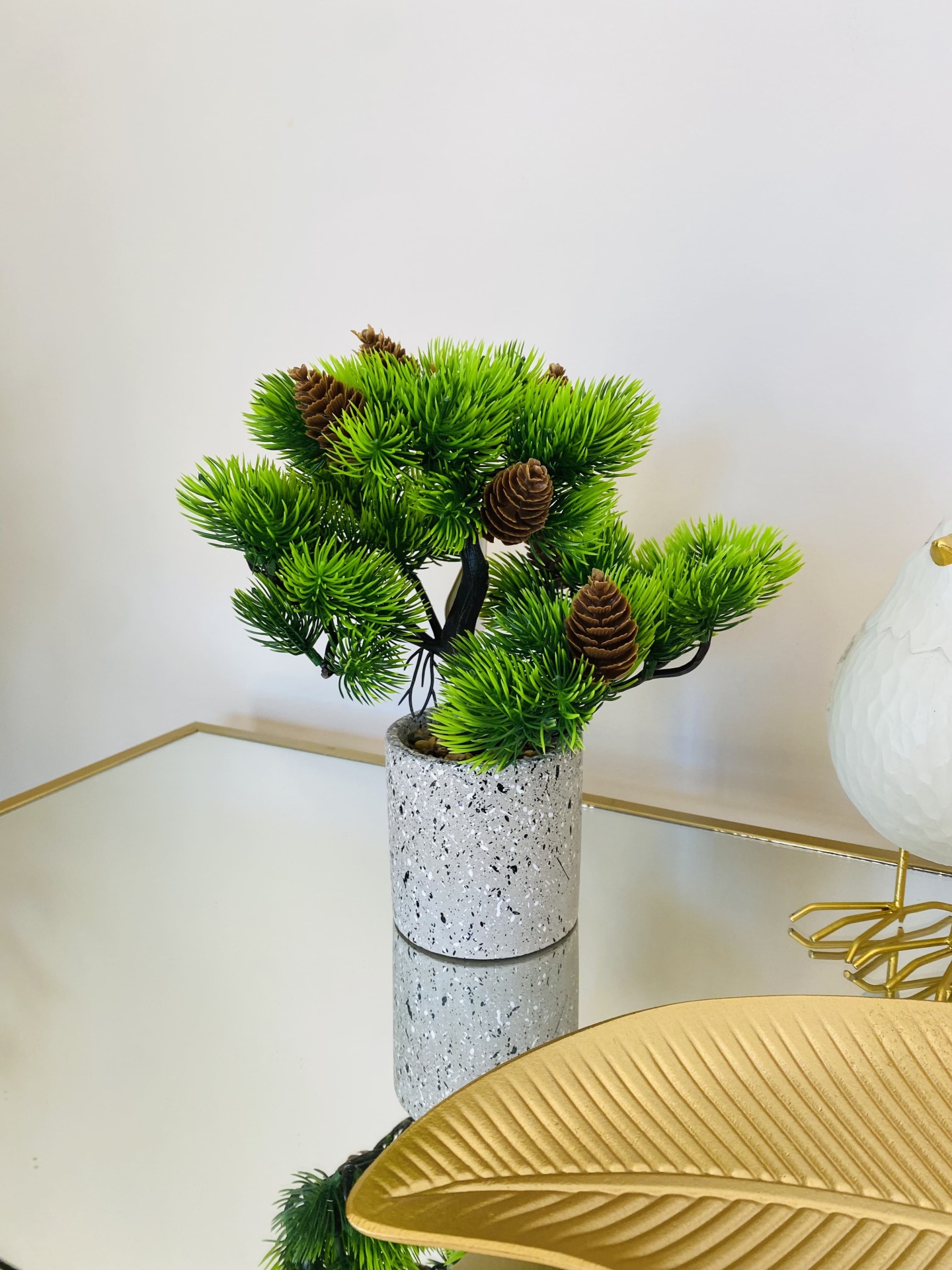 Mini planta artificial em vaso cinza sobre superfície espelhada com decoração dourada.