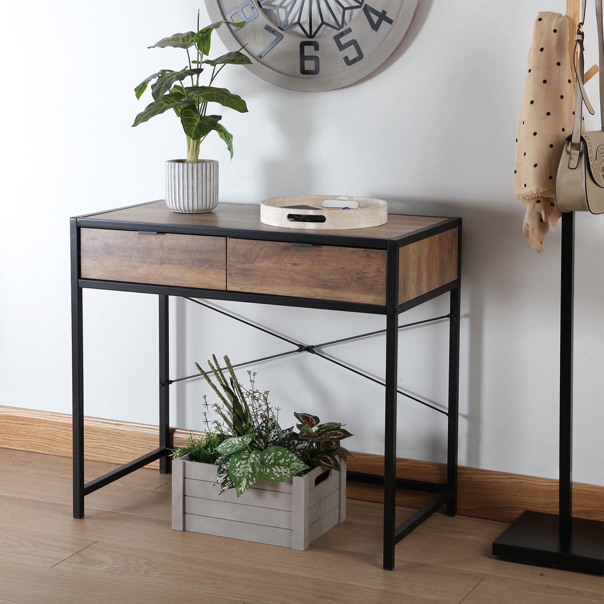 Secretária metálica preta com tampo de madeira castanha e dois gavetões, decorada com planta em vaso e bandeja, com relógio cinzento e suporte de roupa no fundo
