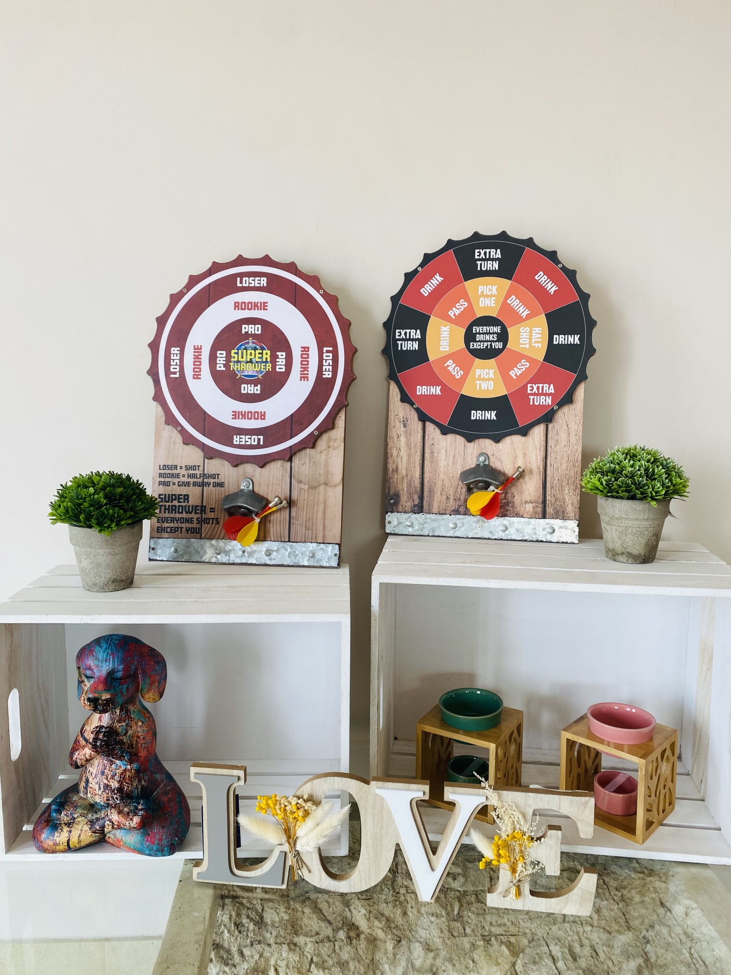 Dois jogos de dardos decorativos com base de madeira e ganchos metálicos sobre caixas brancas com plantas e objetos decorativos