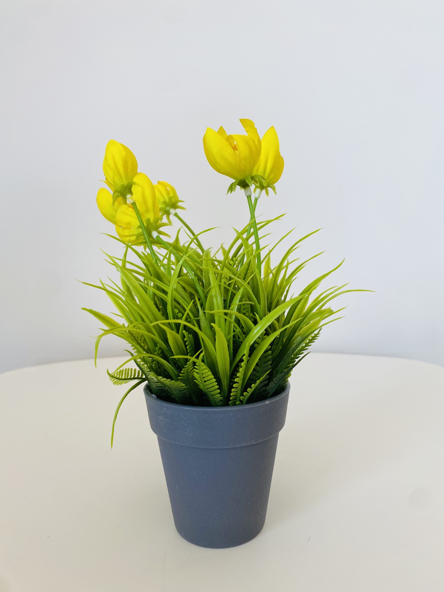 Vaso cinzento com planta artificial verde e flores amarelas sobre superfície branca