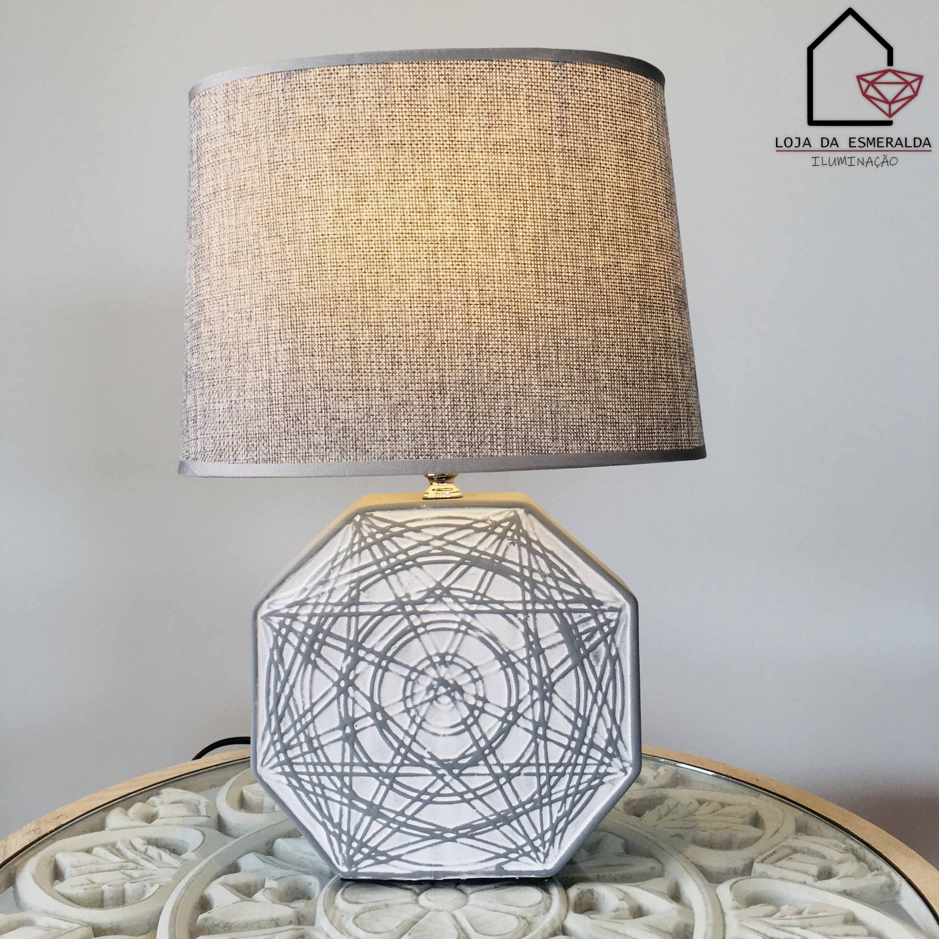 Candeeiro de mesa branco com padrão circular cinzento e cúpula bege em tecido numa mesa redonda com relevo floral.