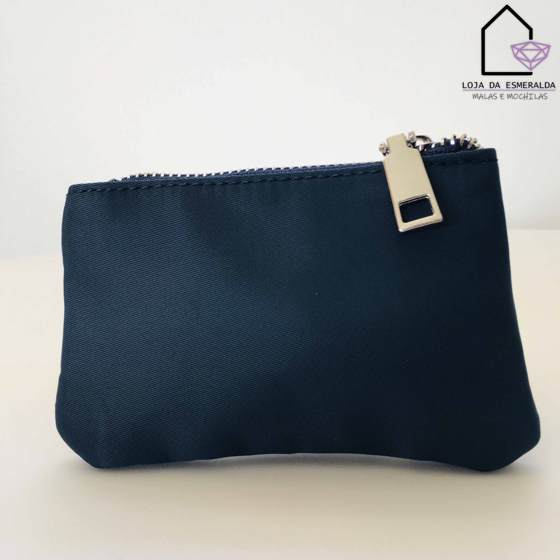 Carteira pequena azul escuro com fecho prateado sobre superfície branca, logo da loja ao fundo