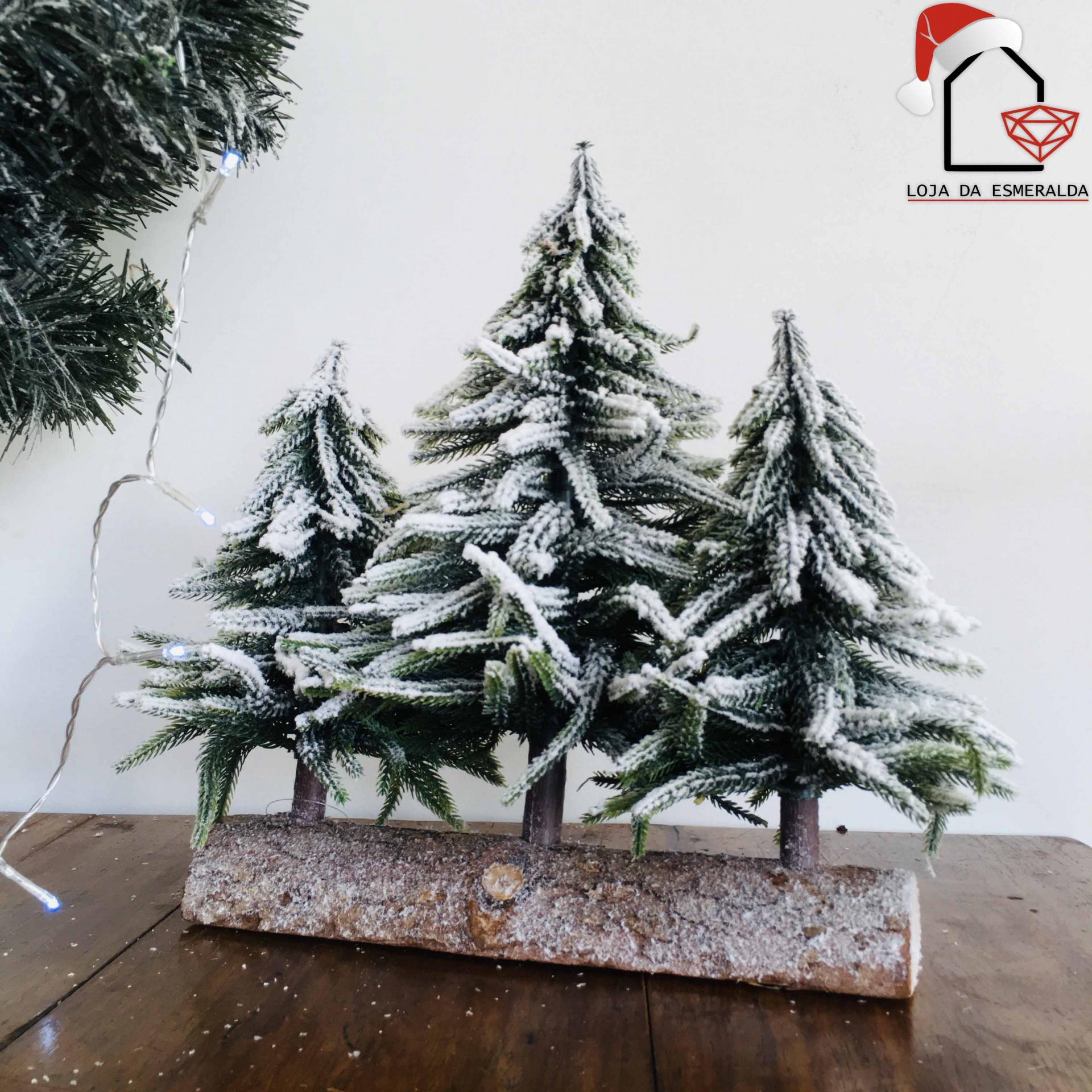 Decoração de Natal com três árvores verdes nevadas numa base de tronco sobre mesa de madeira