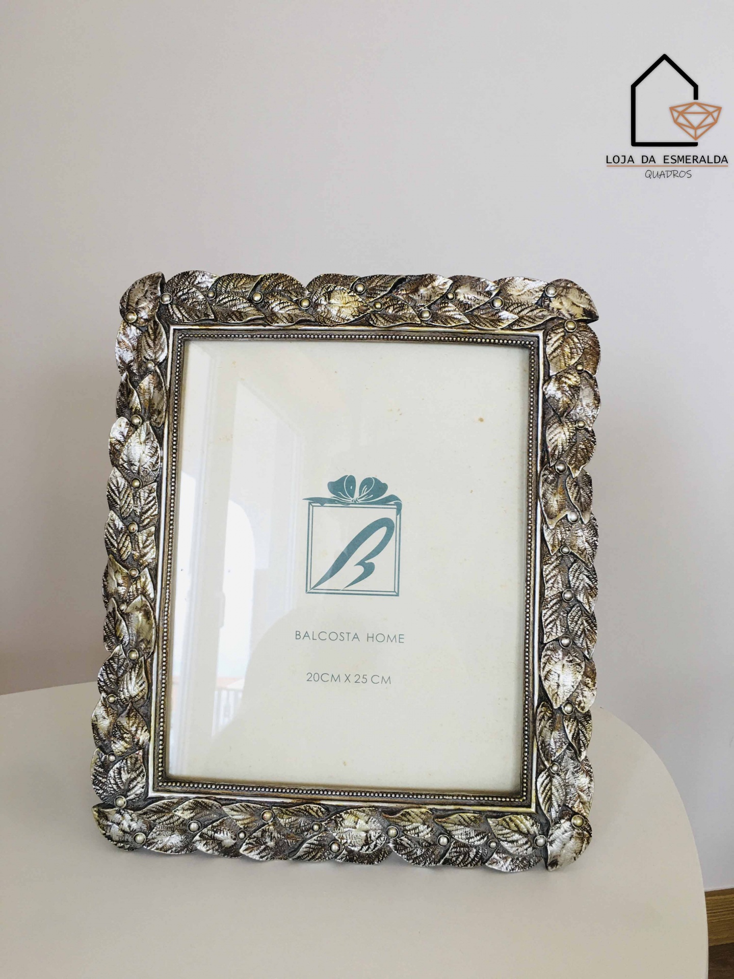 Moldura decorativa com padrão de folhas em metal prateado envelhecido, com logotipo e texto Balcosta Home 20CM X 25 CM.