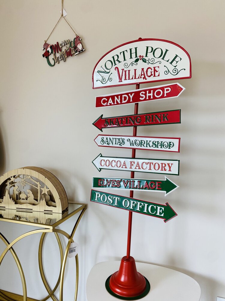 Placa decorativa de Natal com setas indicativas e textos em inglês, suporte vermelho, em mesa branca junto a consola dourada e parede clara.