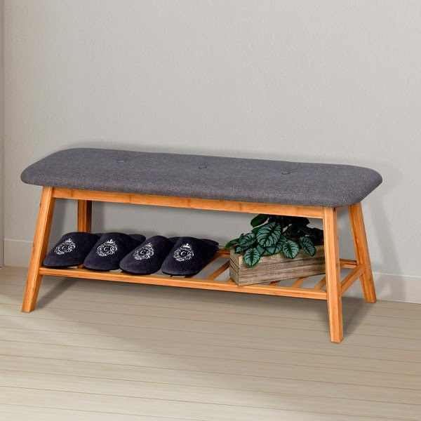 Banco de madeira com assento estofado cinzento, chinelos pretos e planta num vaso de madeira.