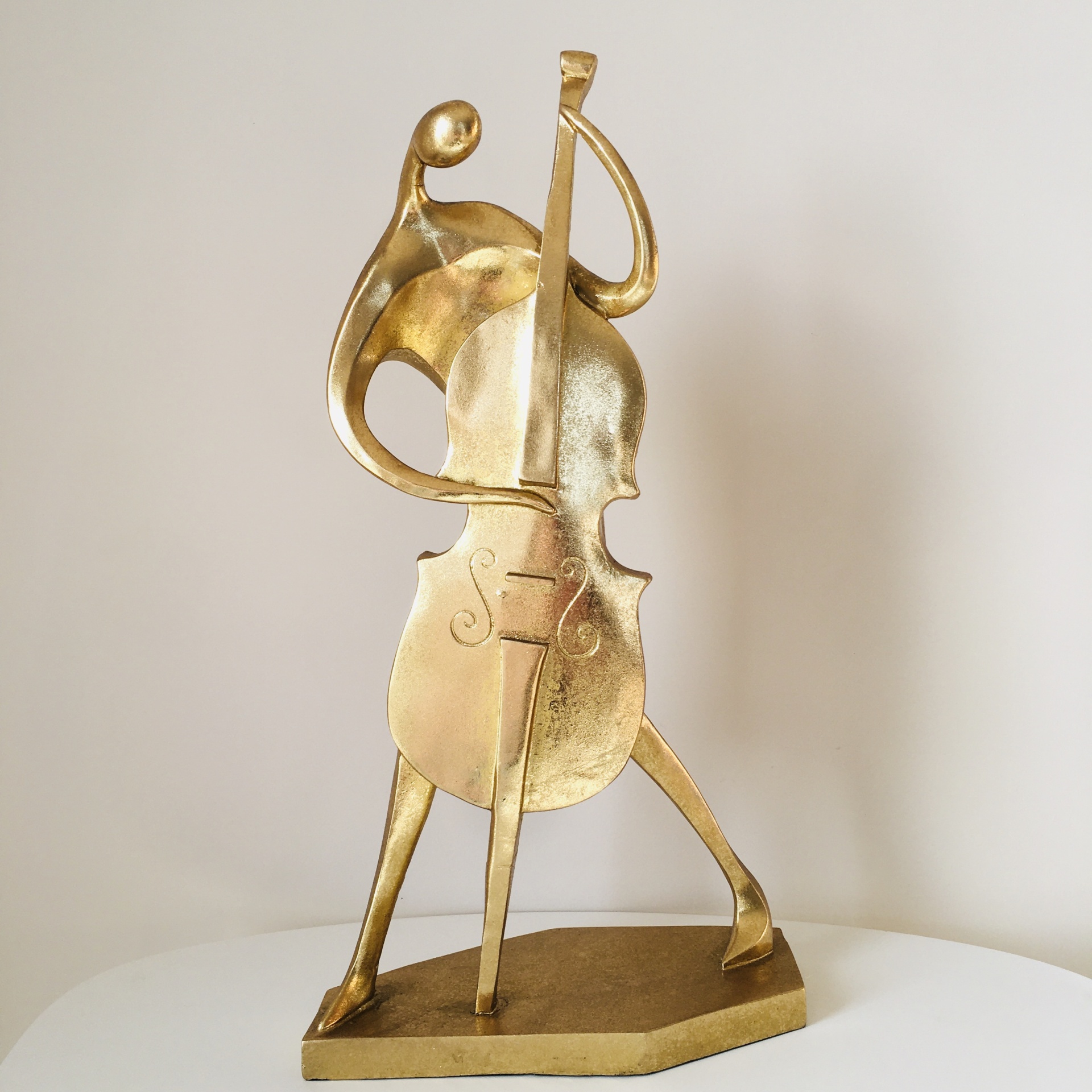 Escultura dourada de figura humana tocando violoncelo sobre base hexagonal