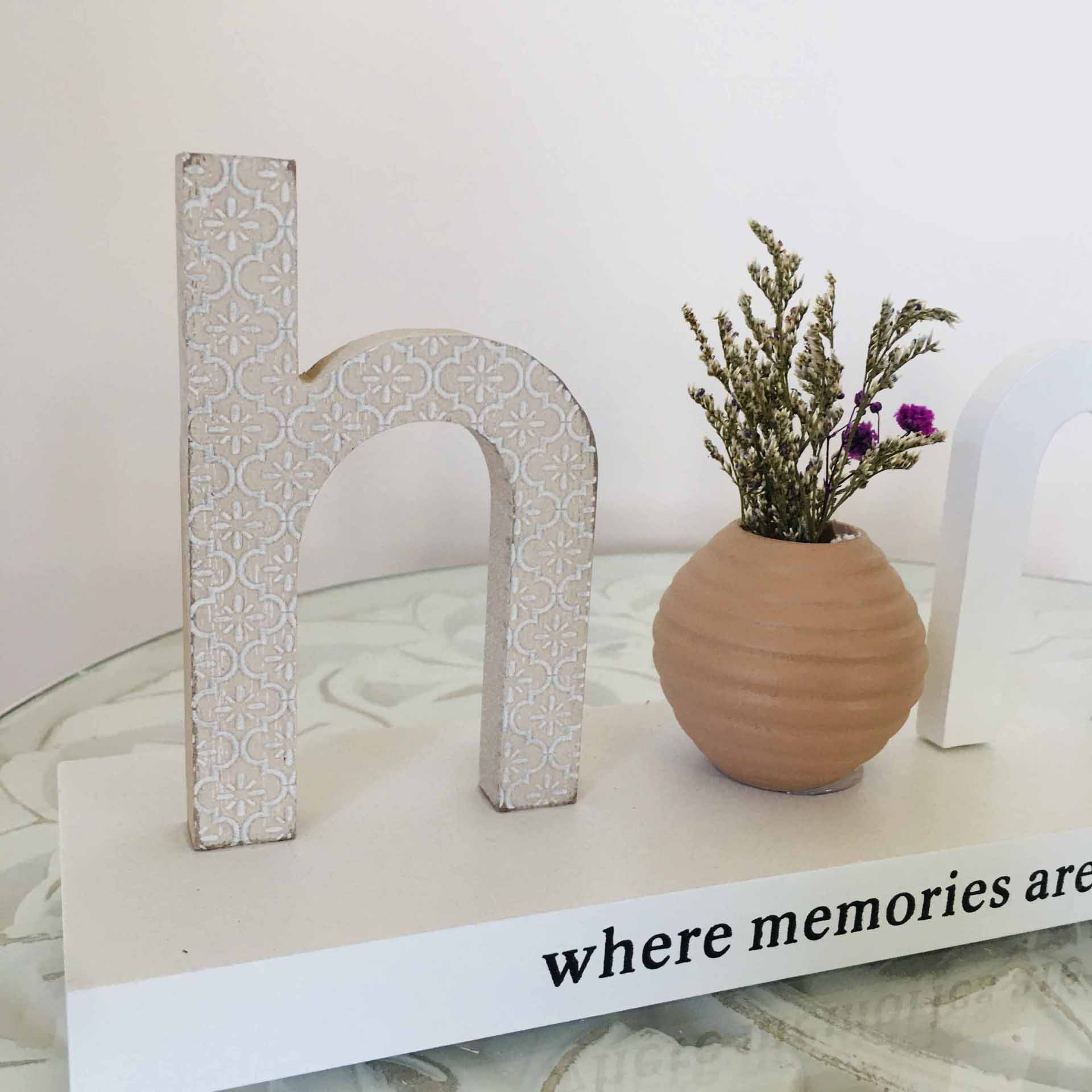 Objeto decorativo letra 'h' em padrão floral e pequeno vaso de cerâmica com flores secas sobre superfície branca