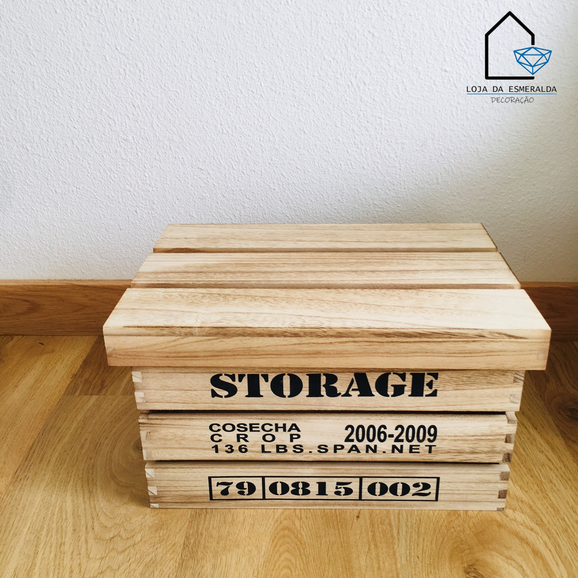 Caixa de madeira clara com tampa e texto preto estampado, sobre chão de madeira e parede branca.