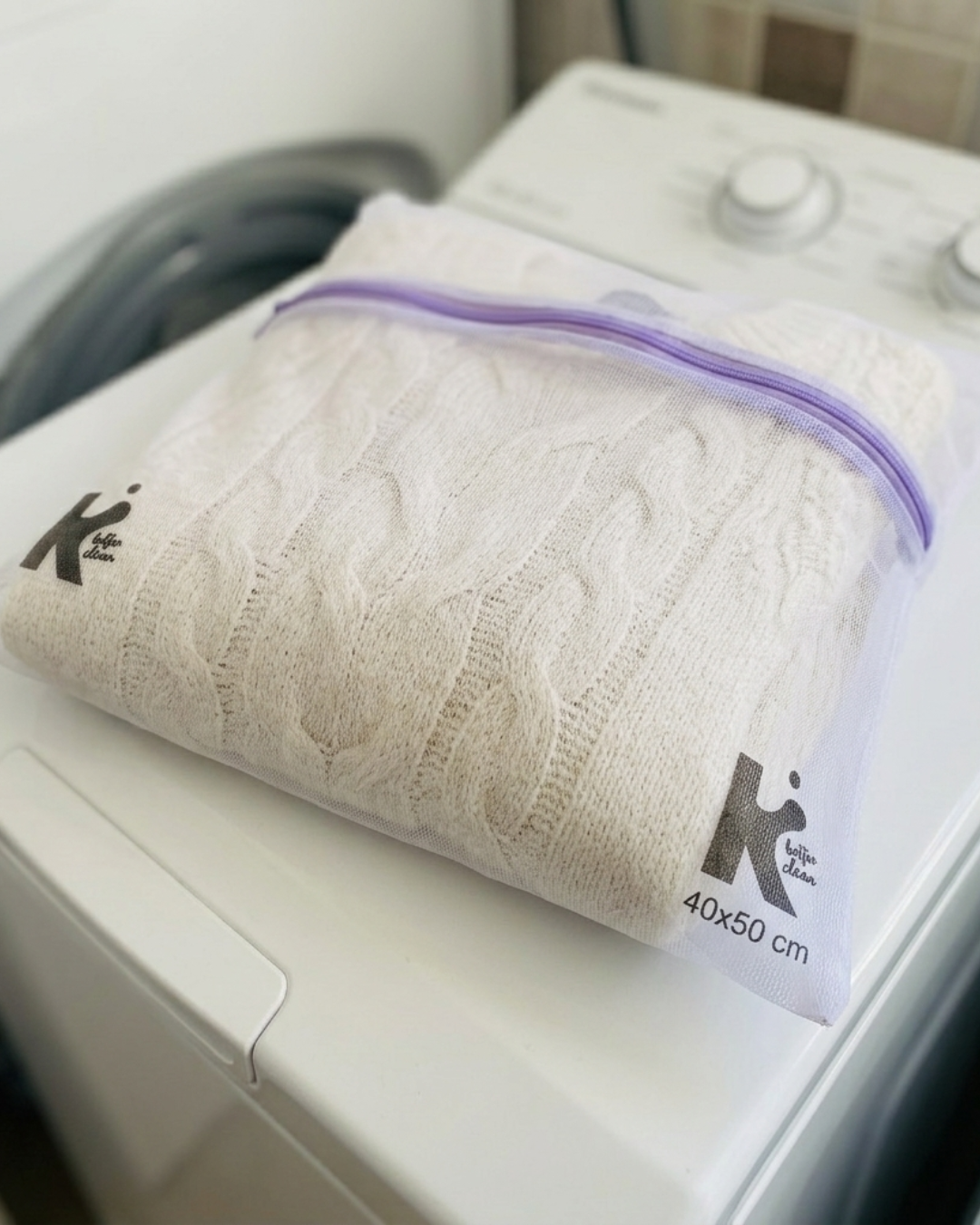 Saco de malha branco com zip violeta e camisola bege dentro, sobre máquina de lavar roupa branca.