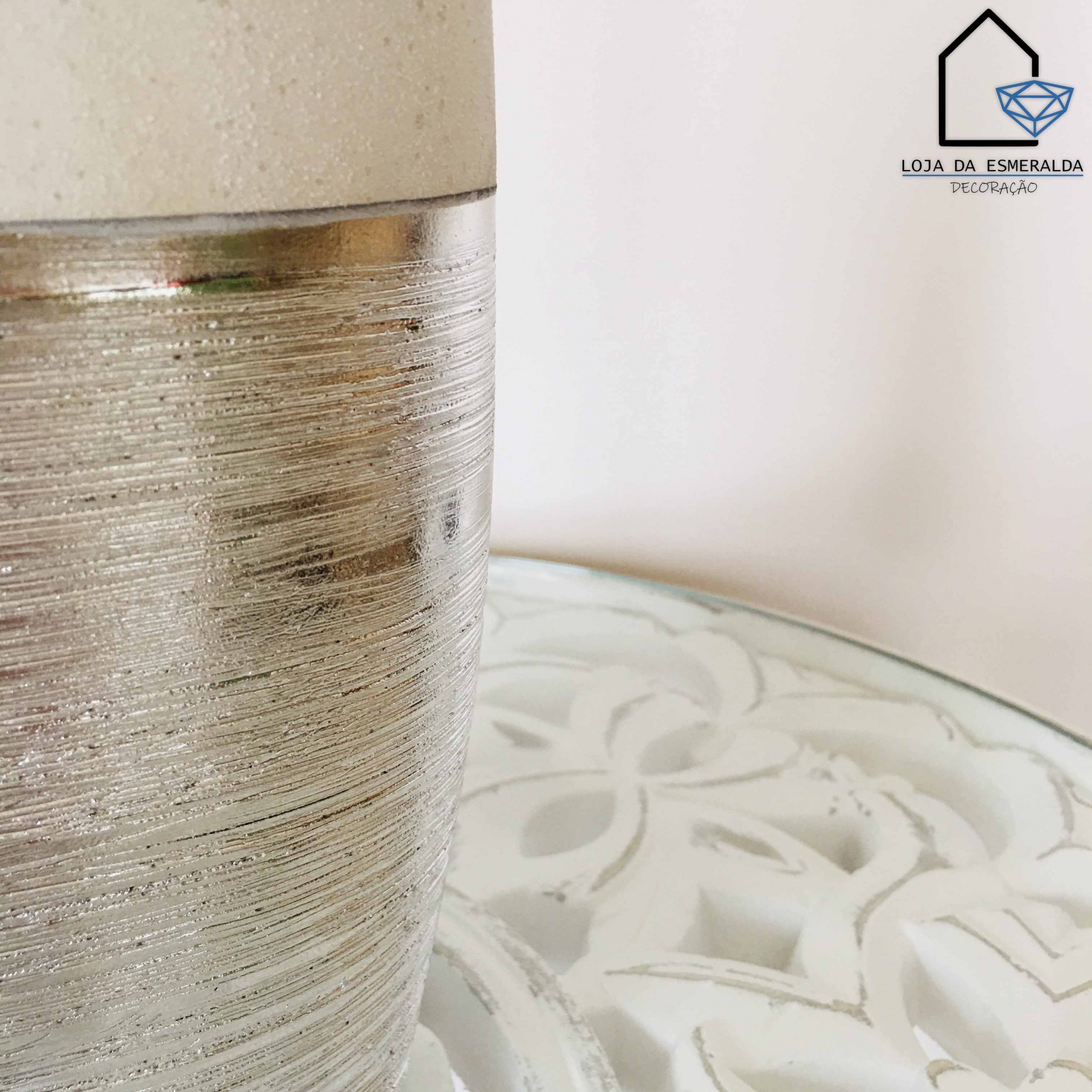 Vaso decorativo prateado e bege sobre mesa branca com padrão floral em relevo.