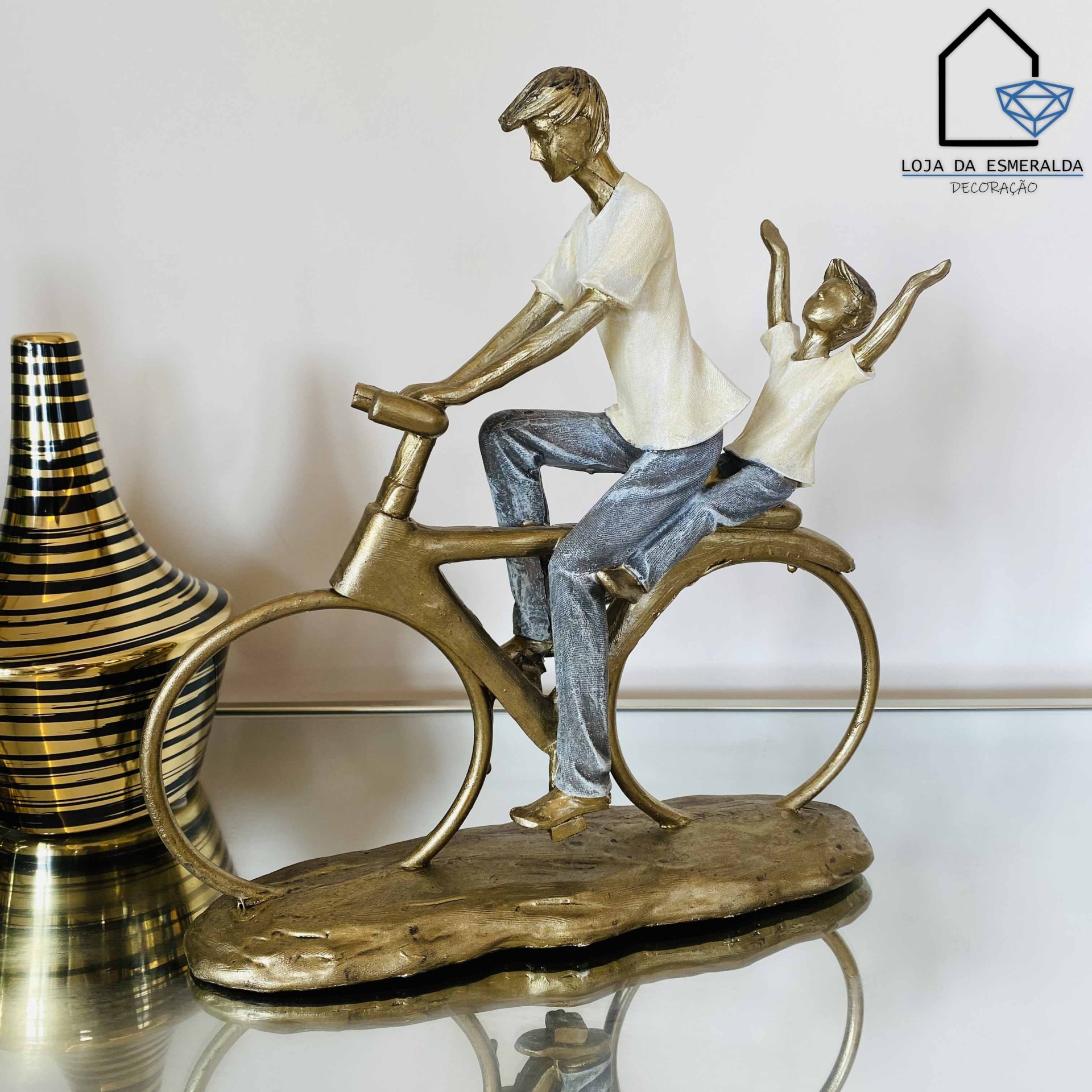 Estatueta de duas figuras humanas numa bicicleta em metal dourado sobre superfície de vidro ao lado de vaso decorativo