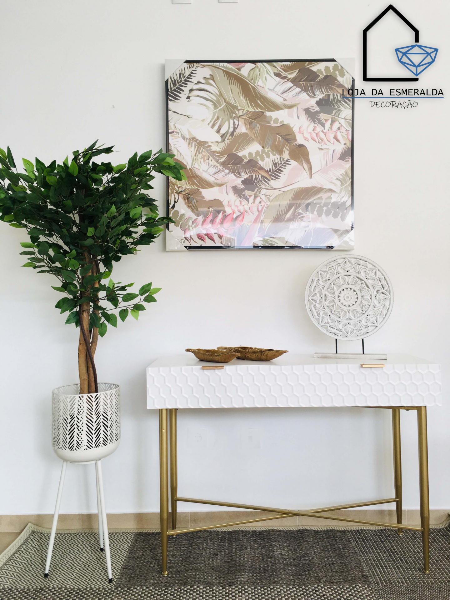 mesa consola branca com padrão hexagonal e pernas douradas, planta artificial em vaso branco, quadro com padrão de folhas e objeto decorativo redondo branco