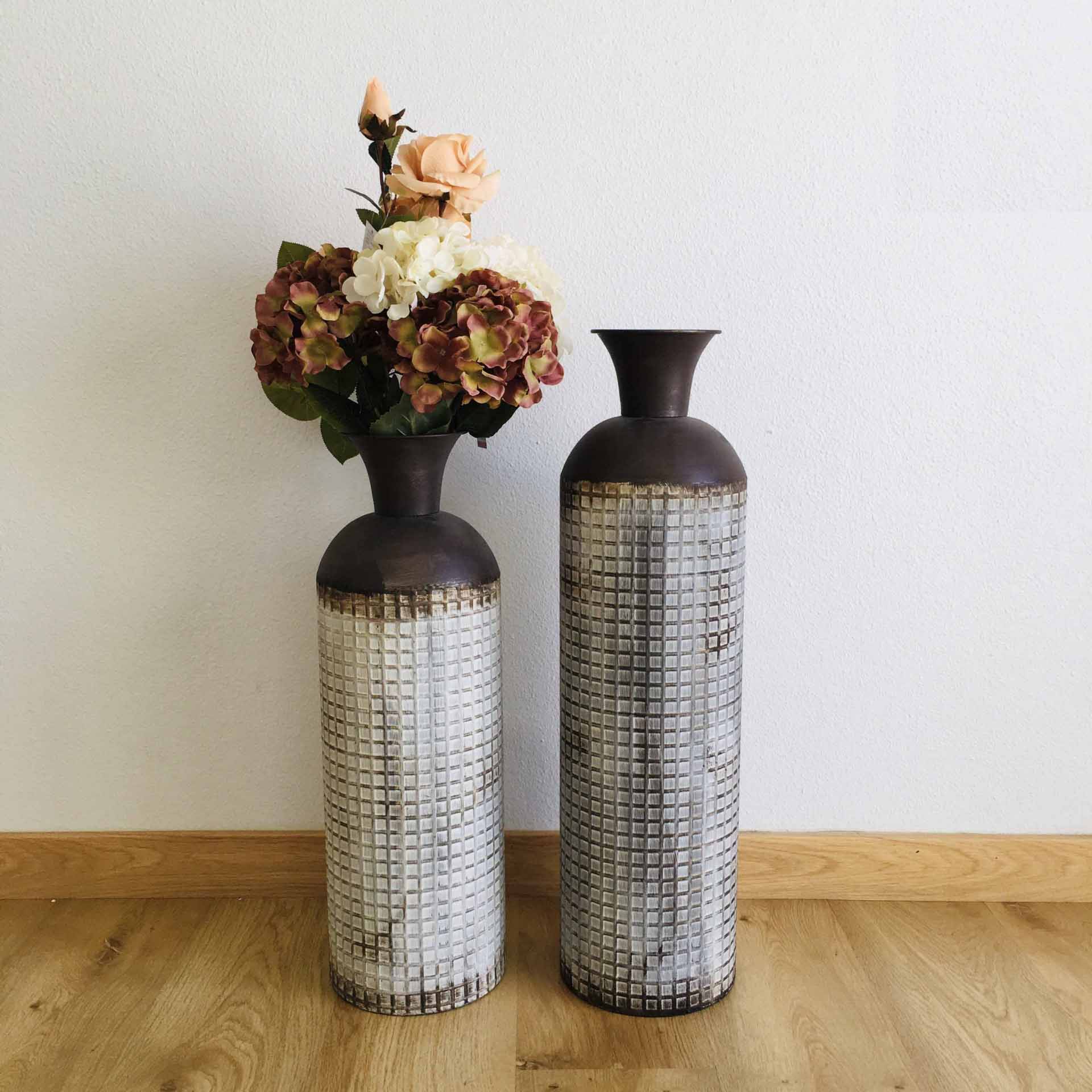Dois vasos decorativos altos em mosaico cinza com topo castanho, um com flores artificiais, sobre chão de madeira junto a parede branca.