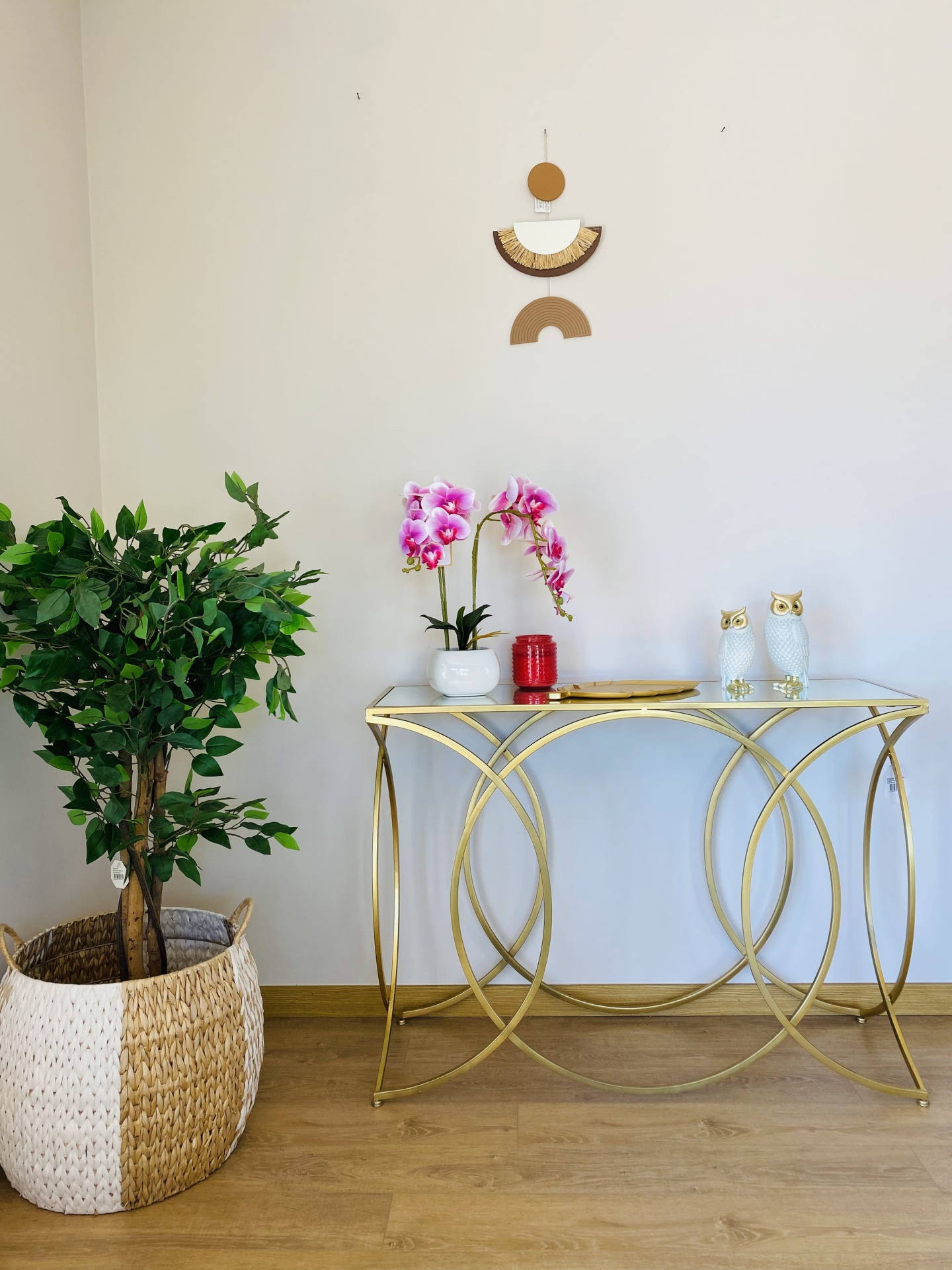 Mesa de apoio dourada com tampo de vidro com orquídeas rosas, corujas brancas e planta verde em cesto ao lado.