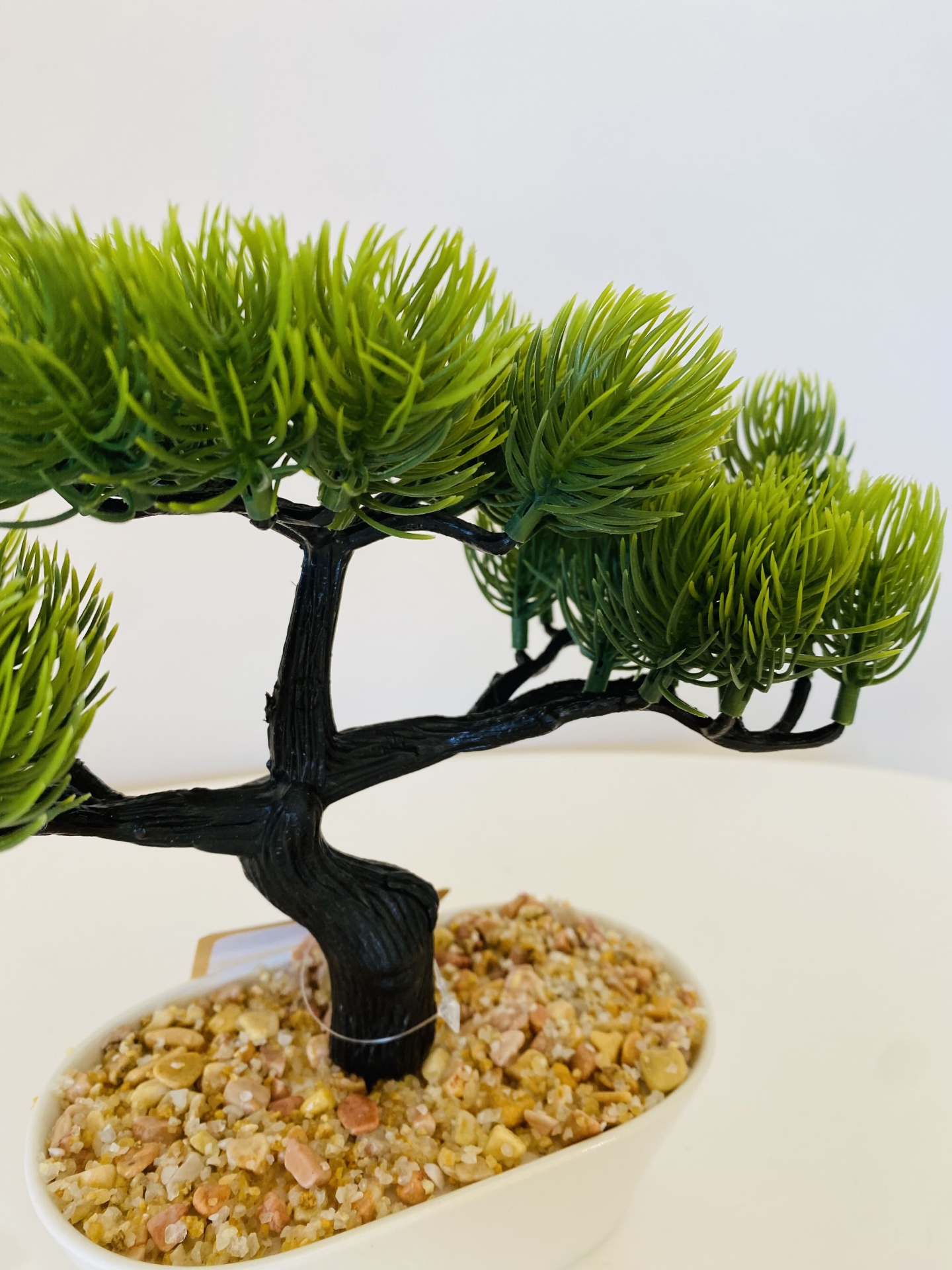 Árvore de bonsai artificial em vaso branco com folhas verdes e tronco preto.